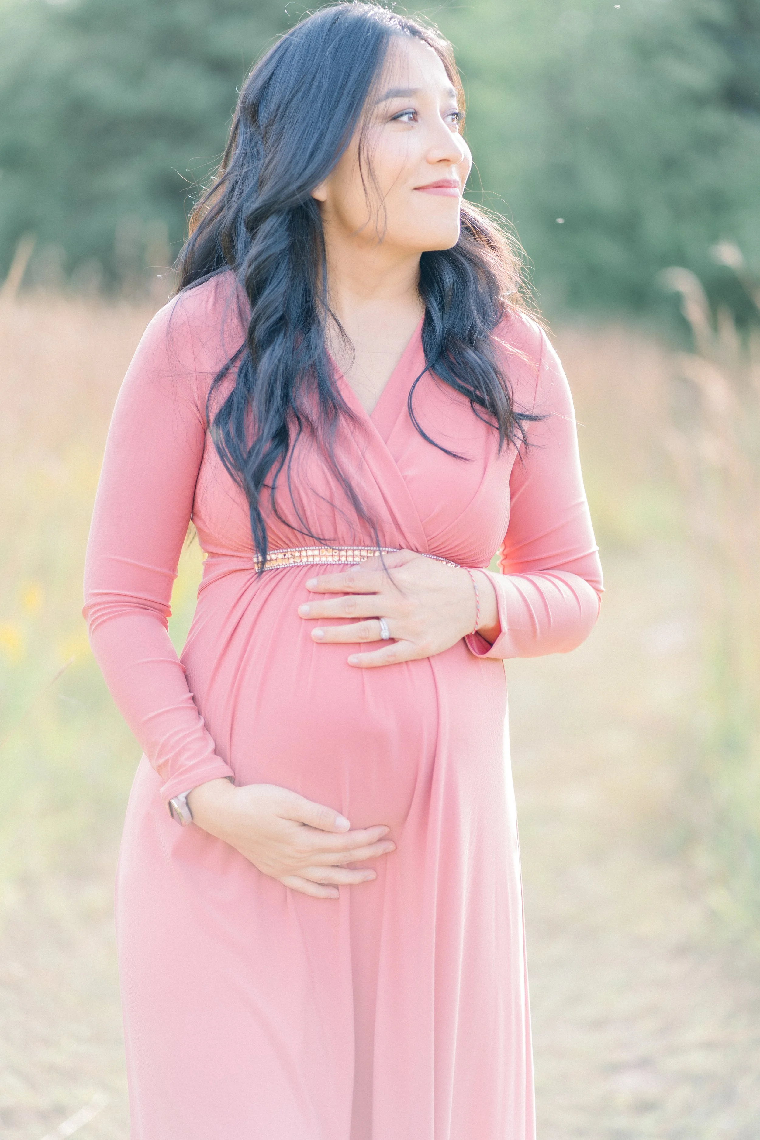 A pregnant woman wearing a long pink dress stands outdoors, gently holding her belly with both hands, and gazes to the right with a soft smile.