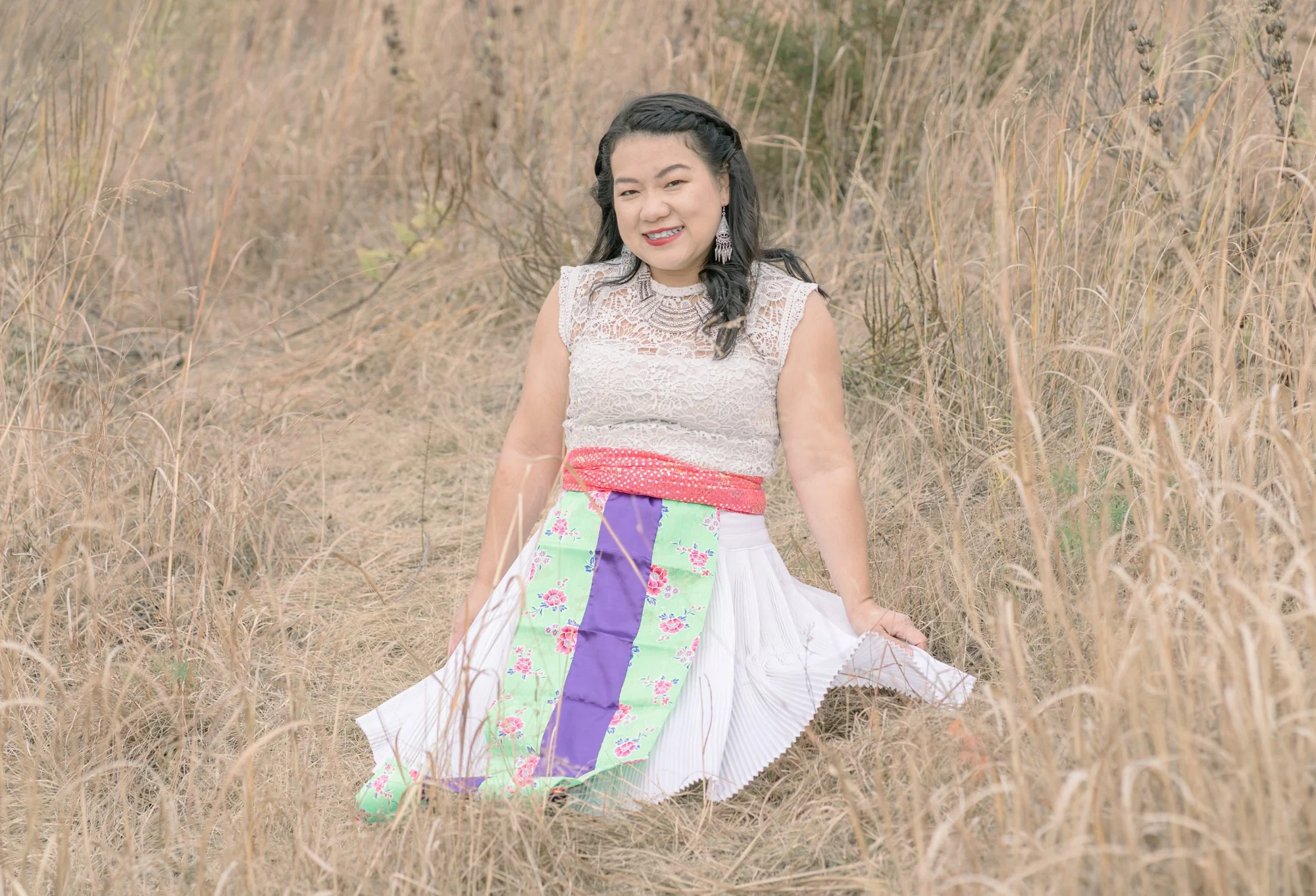 A smiling woman sitting in a field of dry grass, wearing a white lace top, colorful skirt, and silver jewelry.