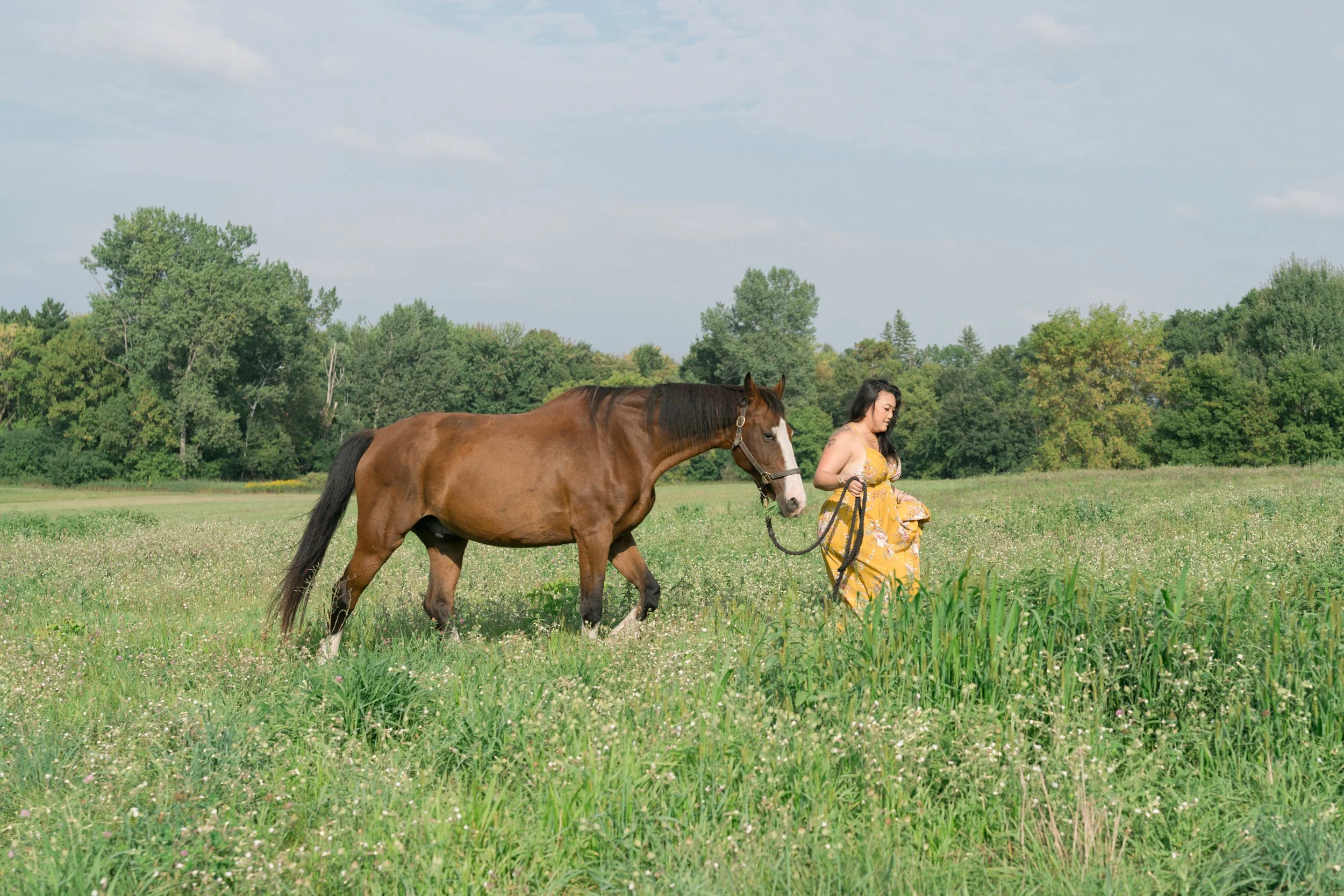 A woman in a yellow dress walking a brown horse through a grassy field with trees in the background under a partly cloudy sky.