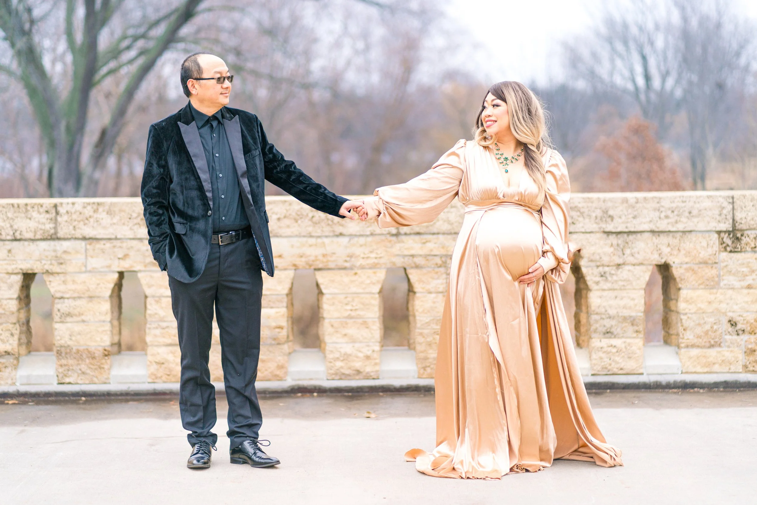 A man and woman standing outdoors on a bridge with trees in the background. The woman, who appears pregnant, is wearing a long, elegant peach-colored dress and a necklace. The man is dressed in a black suit with a black shirt and glasses. They are ho