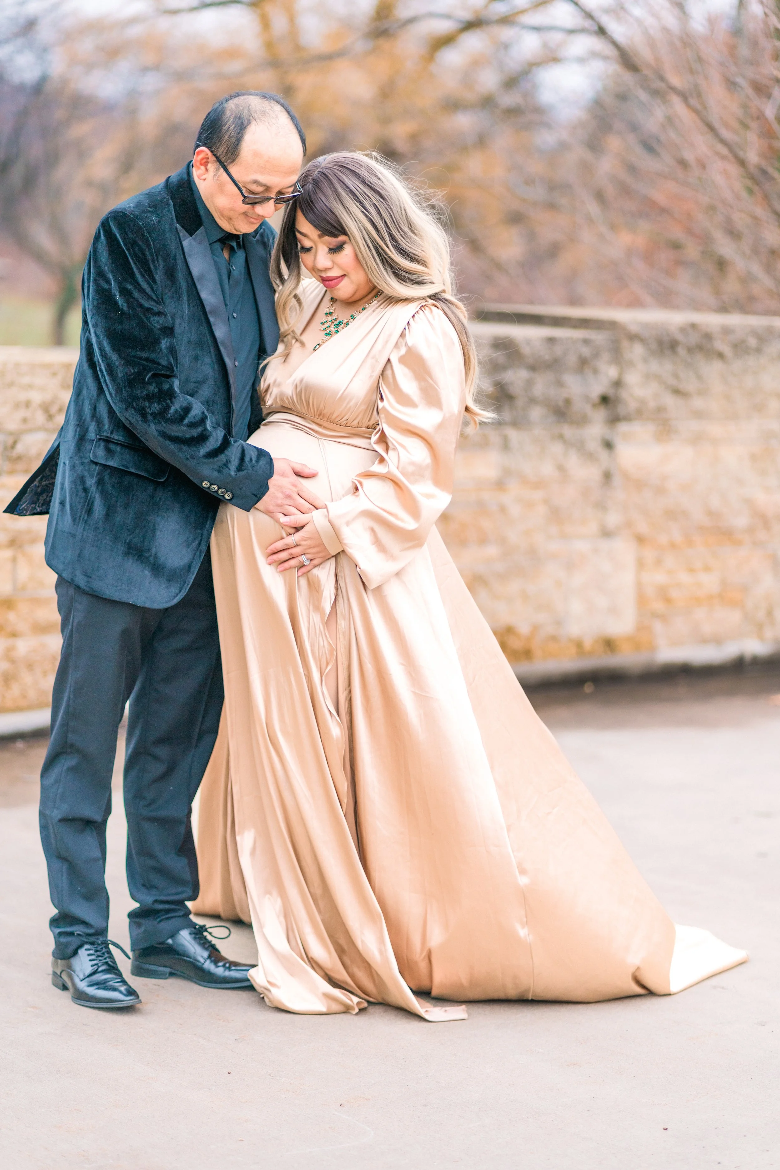 A couple, dressed in formal attire, is standing close together outdoors. The man is touching the woman's pregnant belly as they both look down and smile gently.