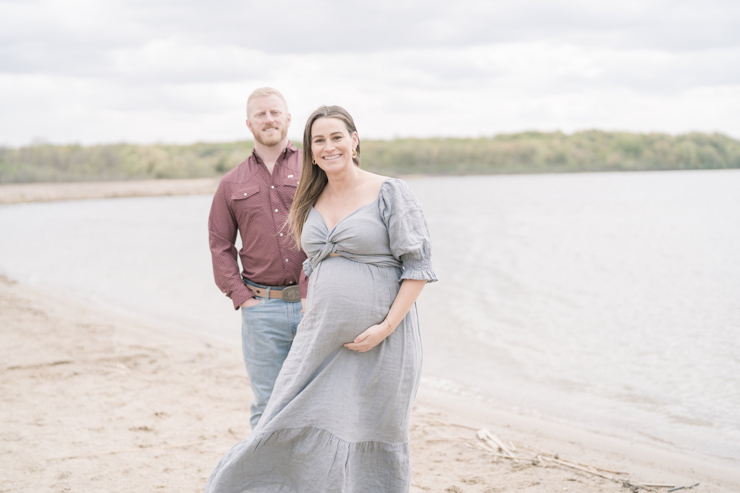 A smiling pregnant woman in a grey dress standing on a sandy beach with a body of water and a treeline in the background, accompanied by a man with a beard and red hair dressed in a maroon shirt and jeans.