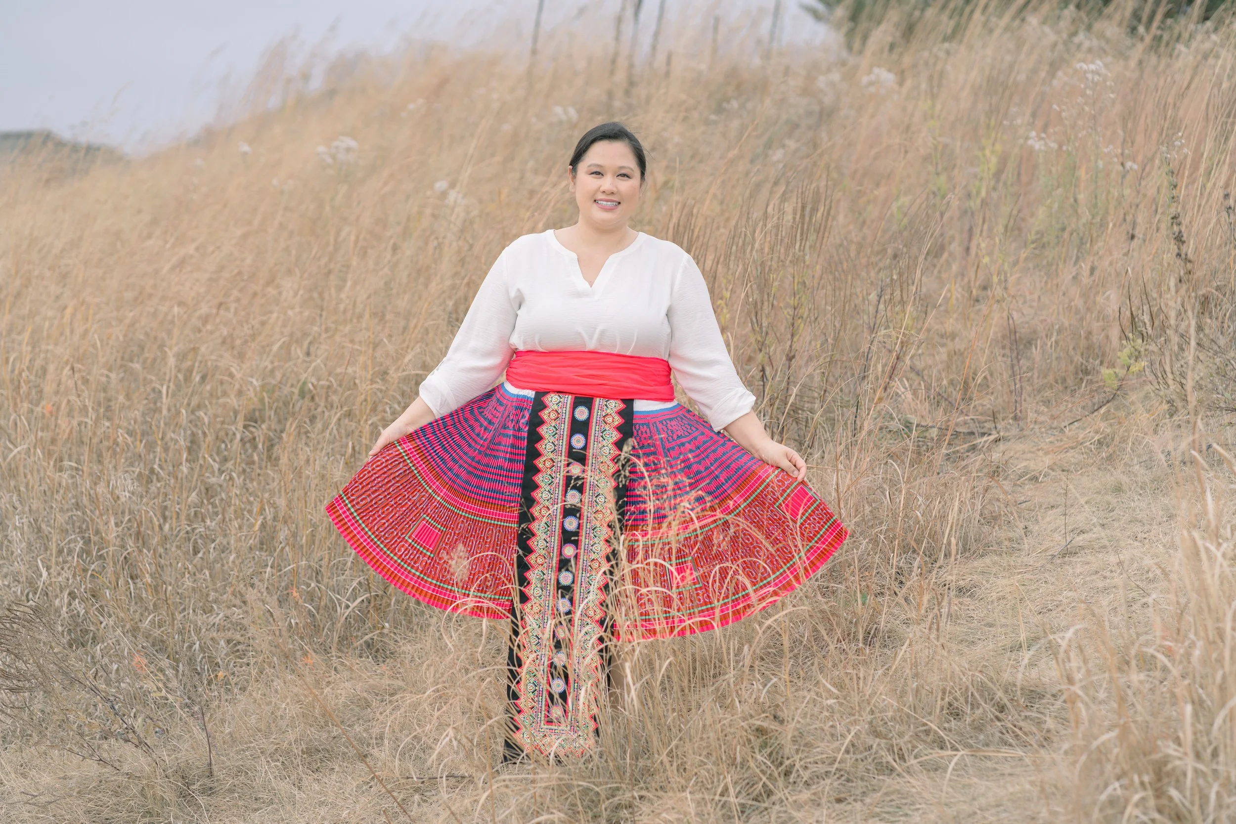 Woman in a white blouse and colorful traditional skirt standing in a dry grassy field.
