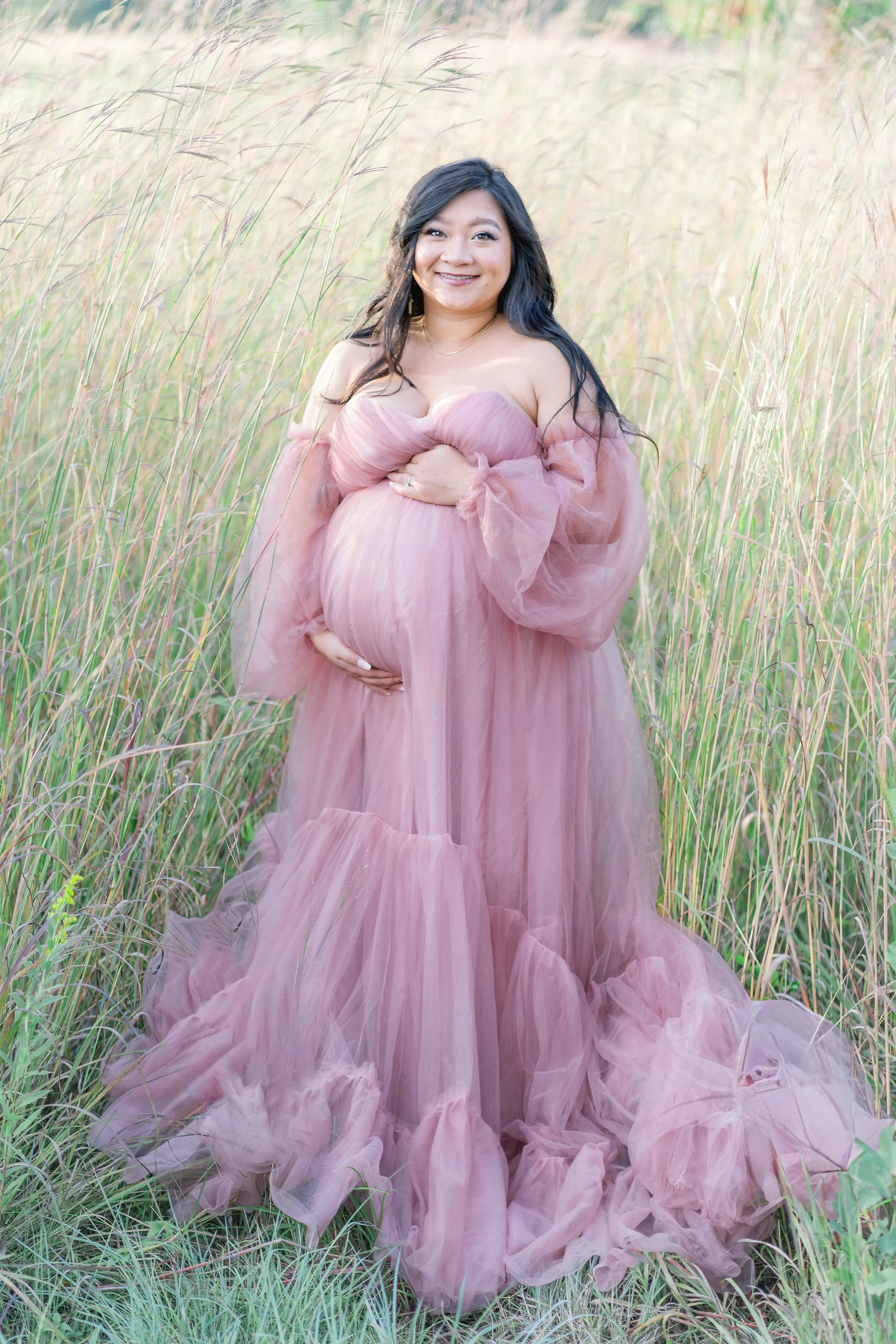 A woman in a flowing pink dress holding her pregnant belly, standing in a field of tall grass with a soft, natural background.