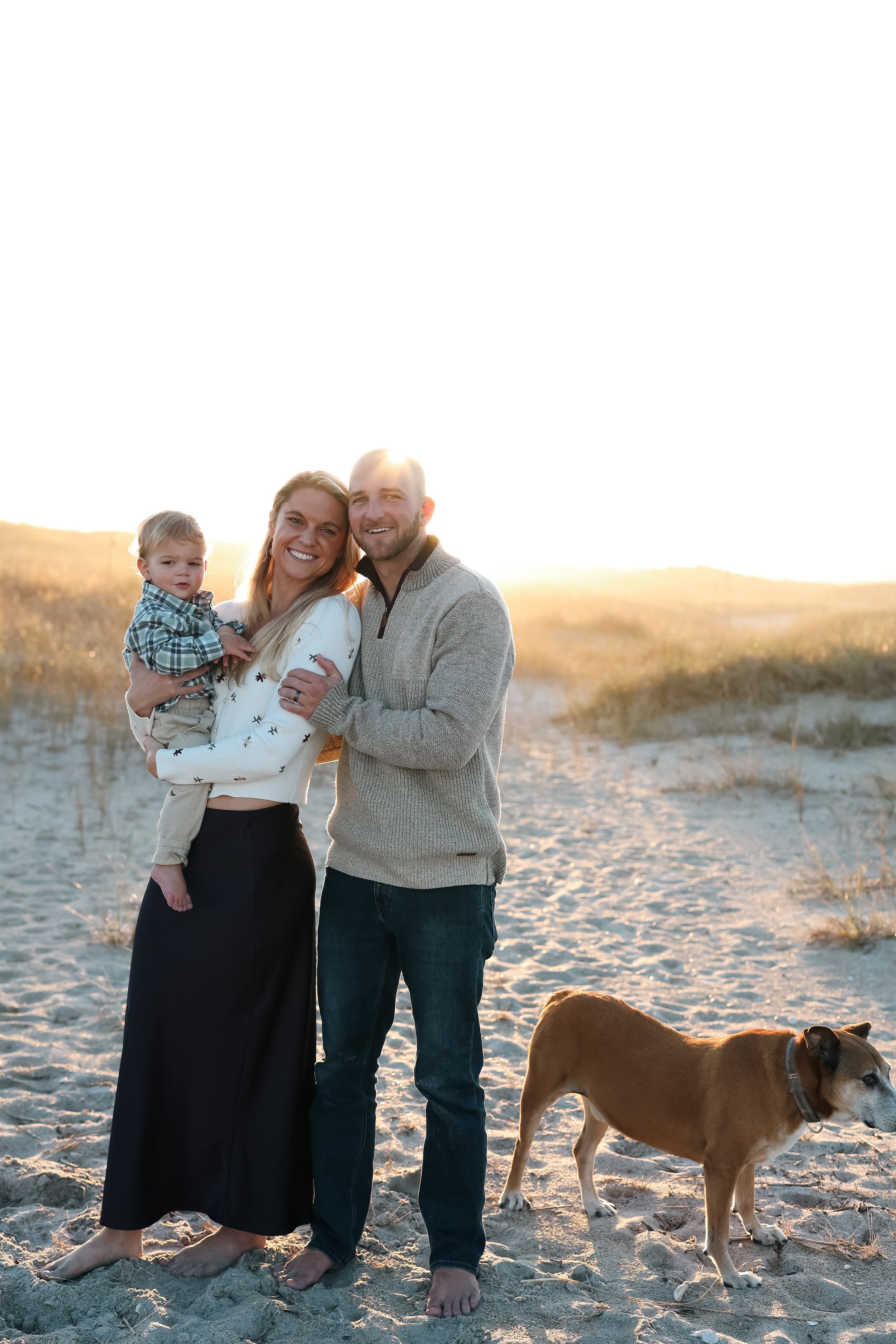 A family of three, with a woman holding a young boy, standing on a beach during sunset, accompanied by a brown dog nearby.