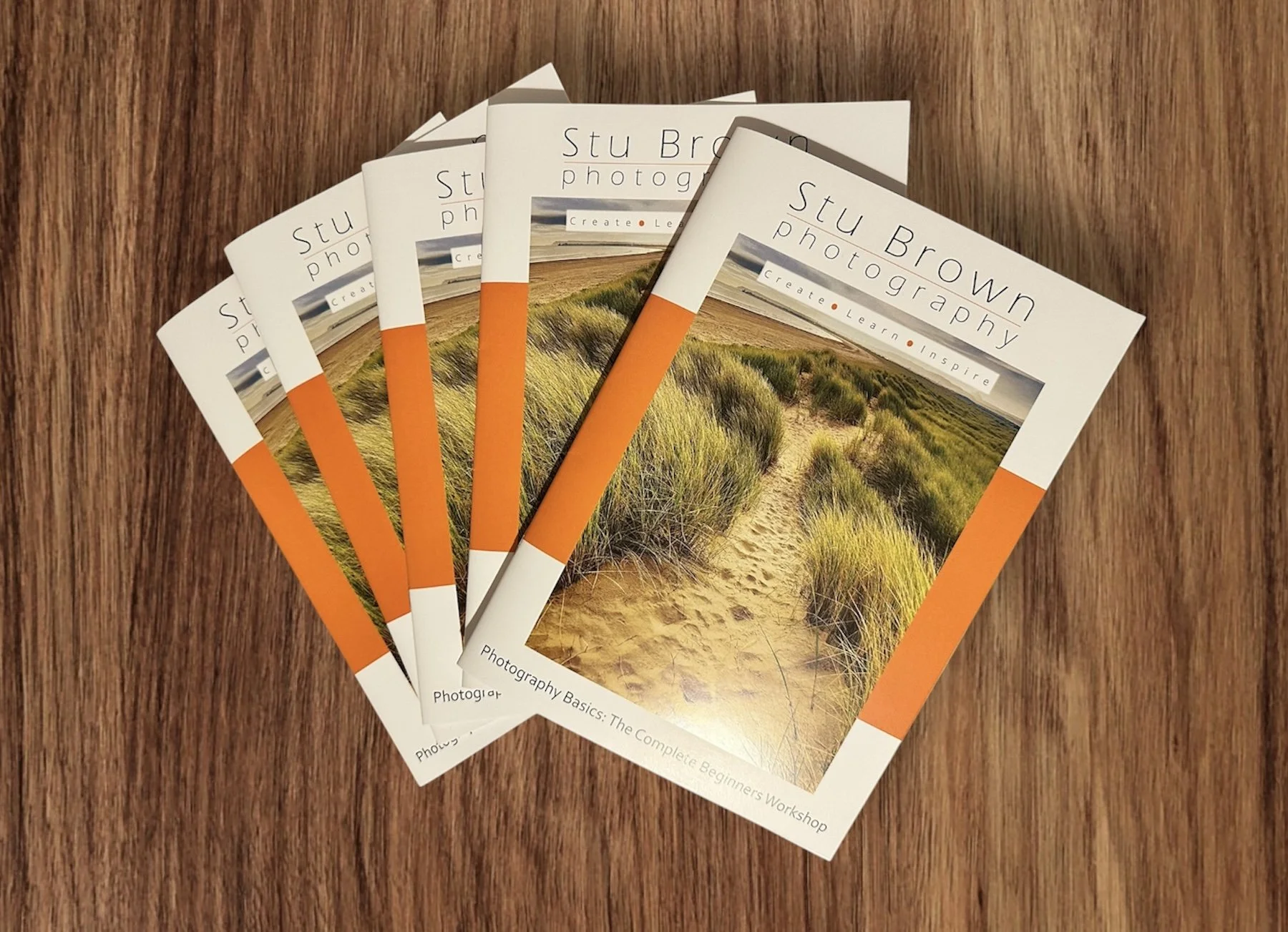 Glossy printed booklets for Stu Brown’s £75 beginner photography workshop, featuring course information and a sand dune landscape cover design, displayed neatly on a wood background.