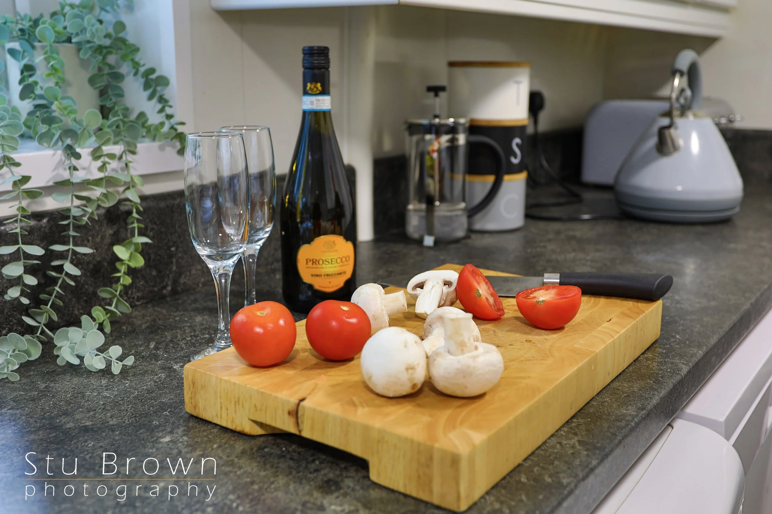 Kitchen countertop with tomatoes, mushrooms, and a knife on a wooden cutting board, a bottle of prosecco, two empty champagne glasses, and kitchen appliances in the background.