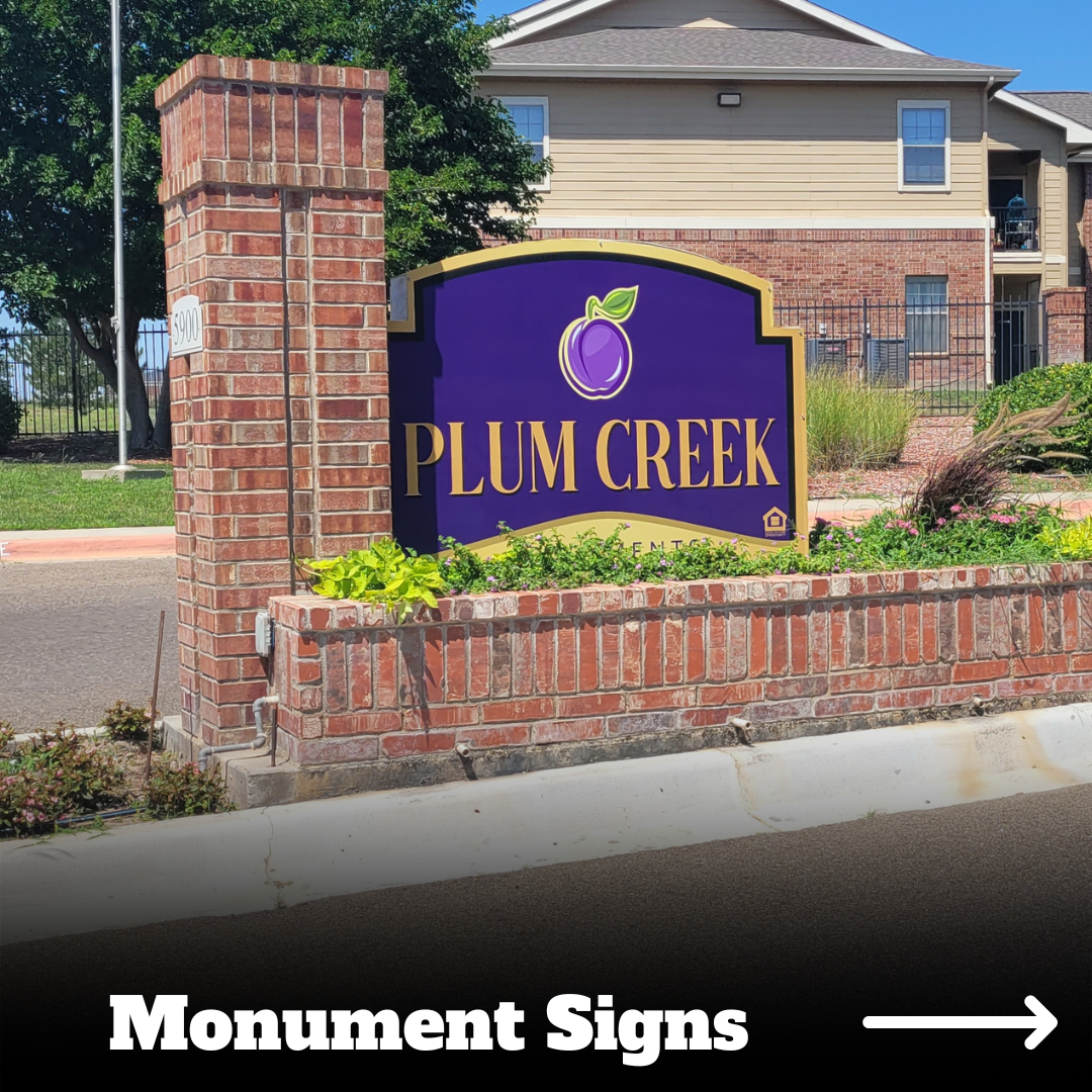A monument sign for Plum Creek Apartments with a purple background, gold border, and a purple plum logo on a brick base surrounded by flowers and greenery, with apartment buildings in the background.