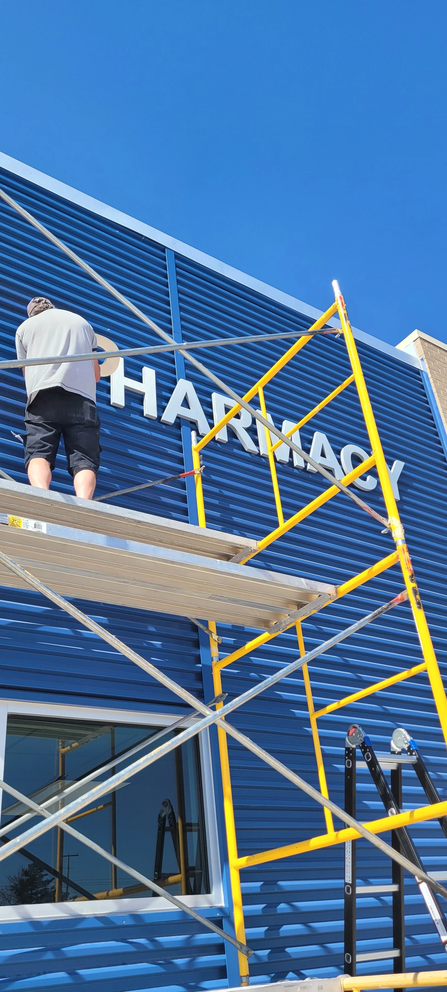 A person on scaffolding installing or repairing a sign on the blue exterior wall of a building, with the partially visible text 'HARMAC'.