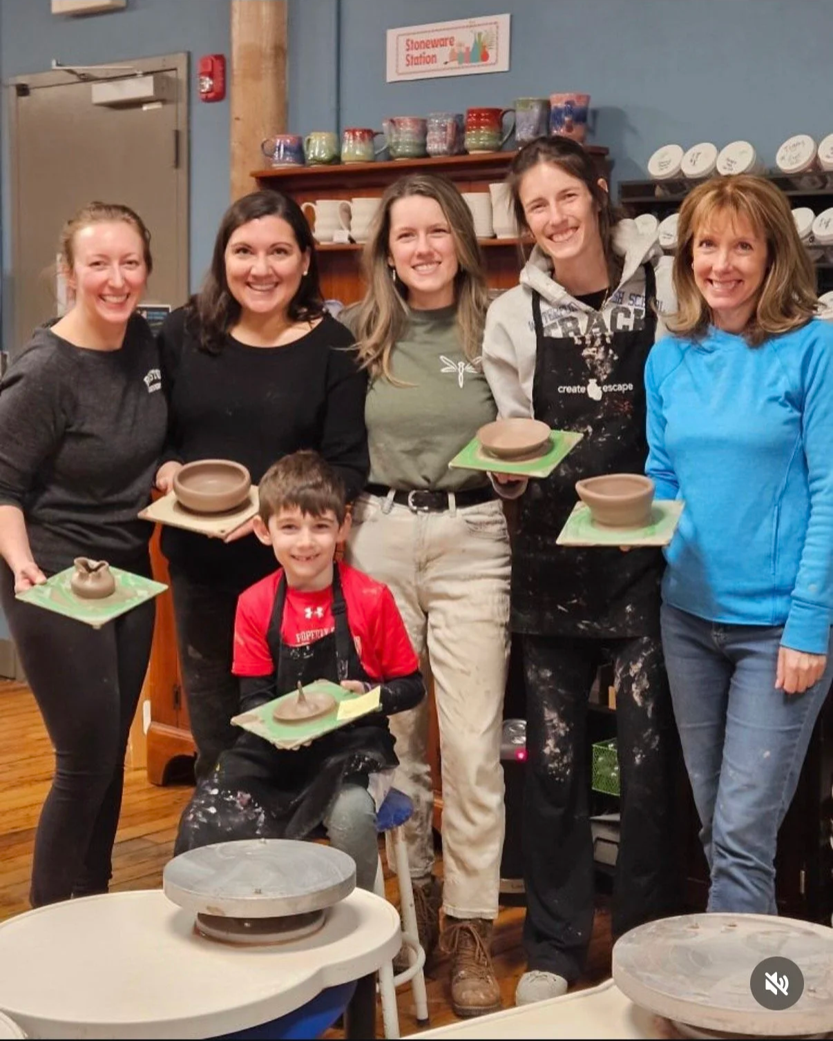 Group of five women and one young boy in a pottery painting class, holding finished and unfinished pottery pieces on trays, with shelves of glazed pottery in the background.
