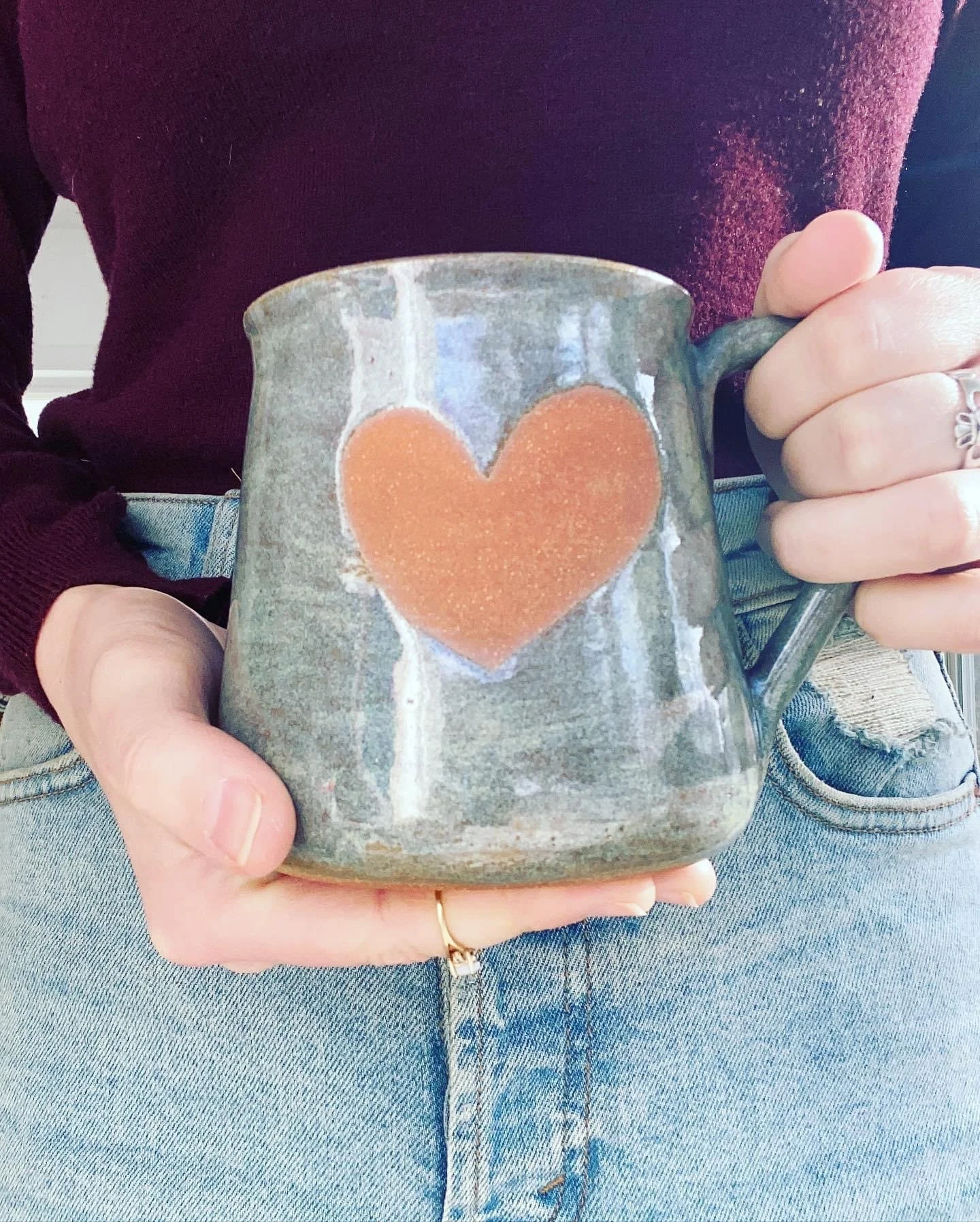 Person holding a ceramic mug with a heart design on it, wearing a maroon top and ripped jeans.