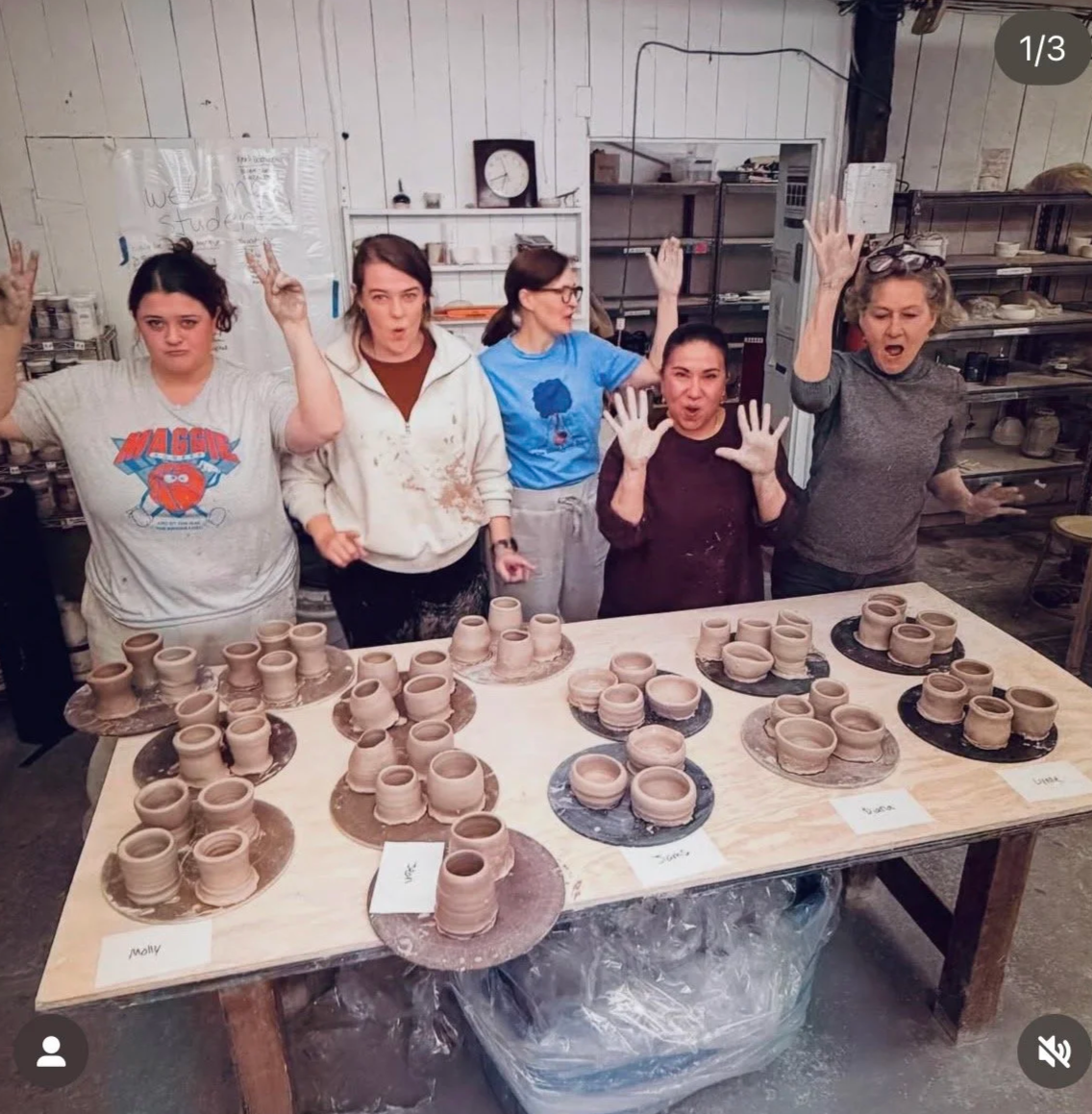 Five women standing behind a table full of unglazed pottery cups, some with small plates, all labeled with names. The women are posing with raised hands, making peace signs or expressive gestures. The setting appears to be a pottery studio with shelves of supplies and a clock on the wall.