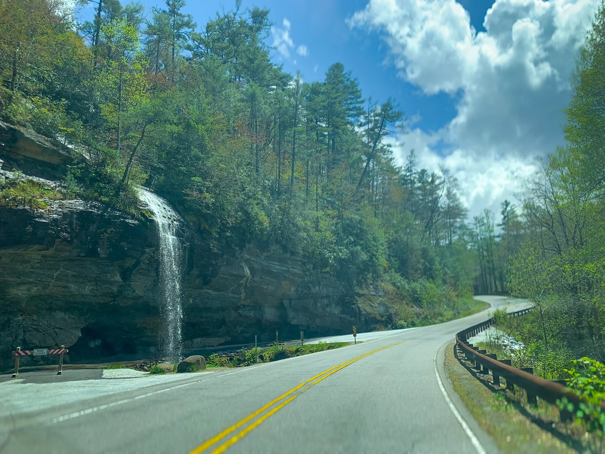 View through a camper van windshield of the steep mountain road passing Bridal Veil Falls near Highlands, North Carolina, with water cascading down a rocky cliff beside the roadway.
