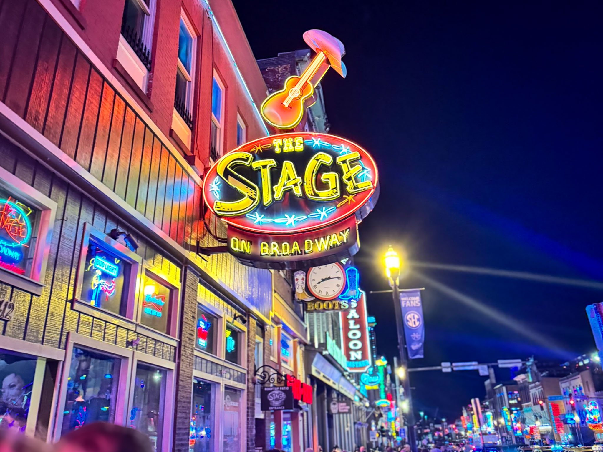 The neon-lit buildings of Lower Broadway in Nashville add a glow to the night sky as crowds stream by on the sidewalk.