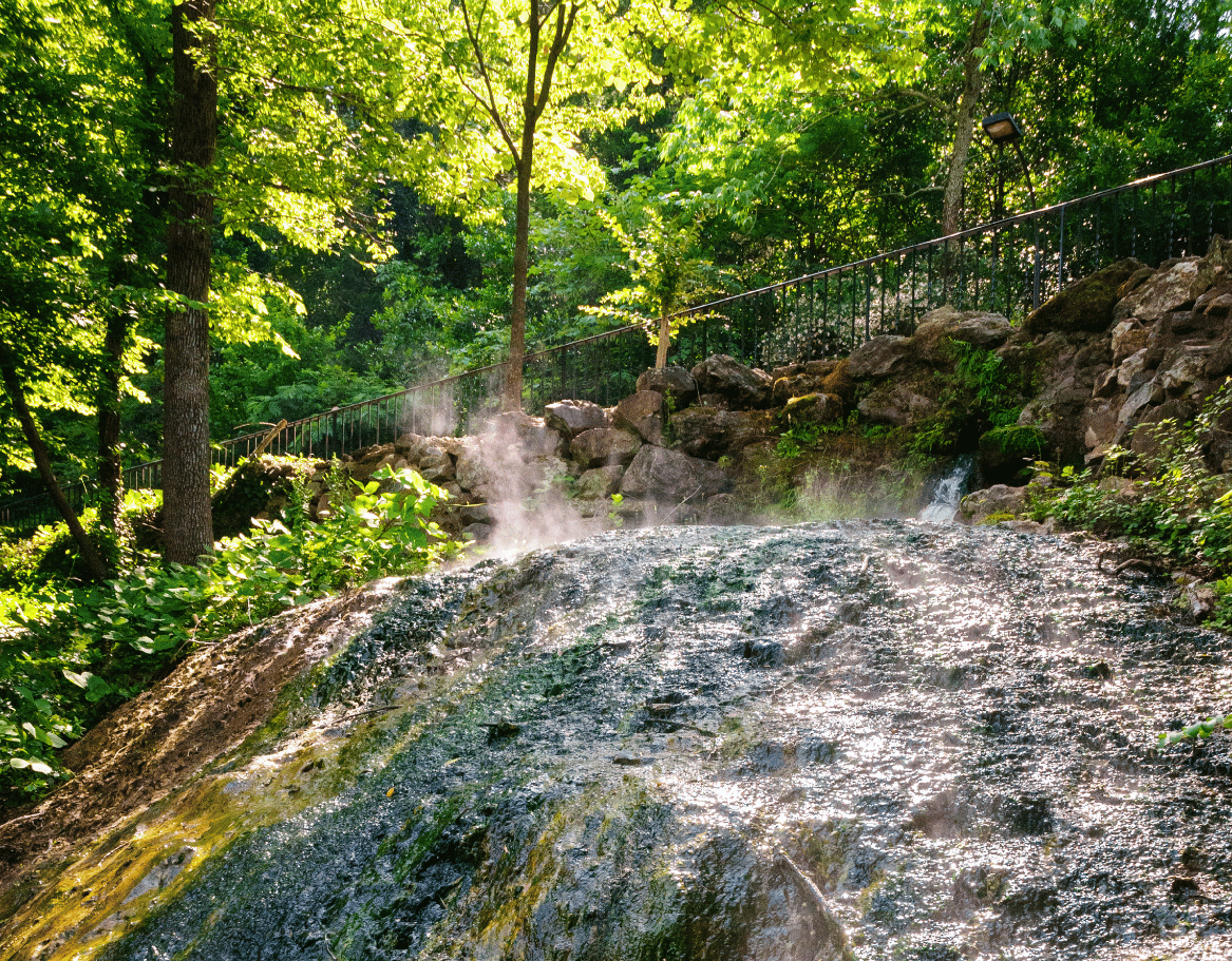Thermal spring water steaming as it flows over rocks surrounded by greenery at Hot Springs National Park, with visible vapor rising from the hot water.