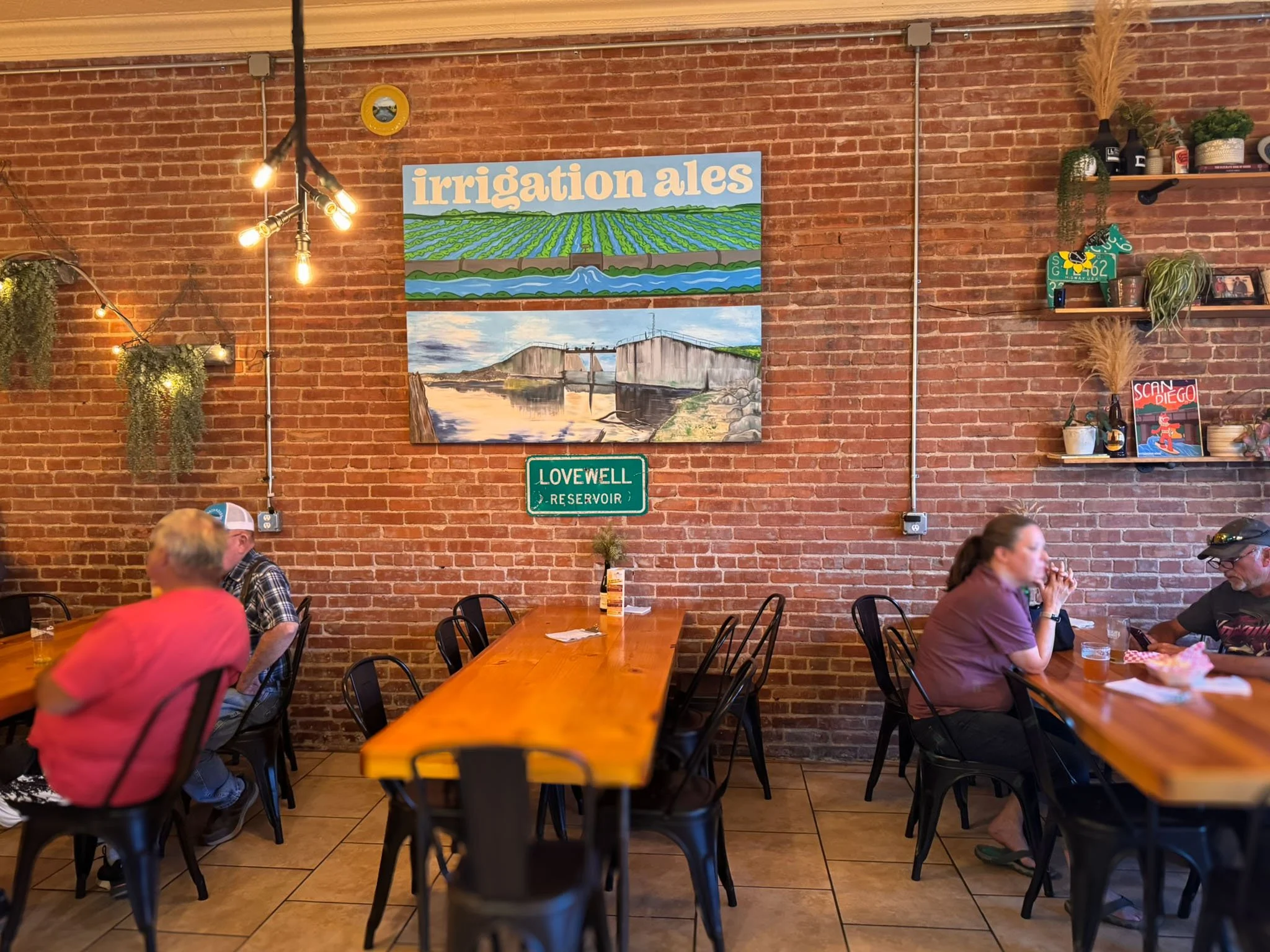 People sit at long, family-style tables inside the taproom of Irrigation Ales, enjoying pints of beer and conversation in a warm, rustic space.