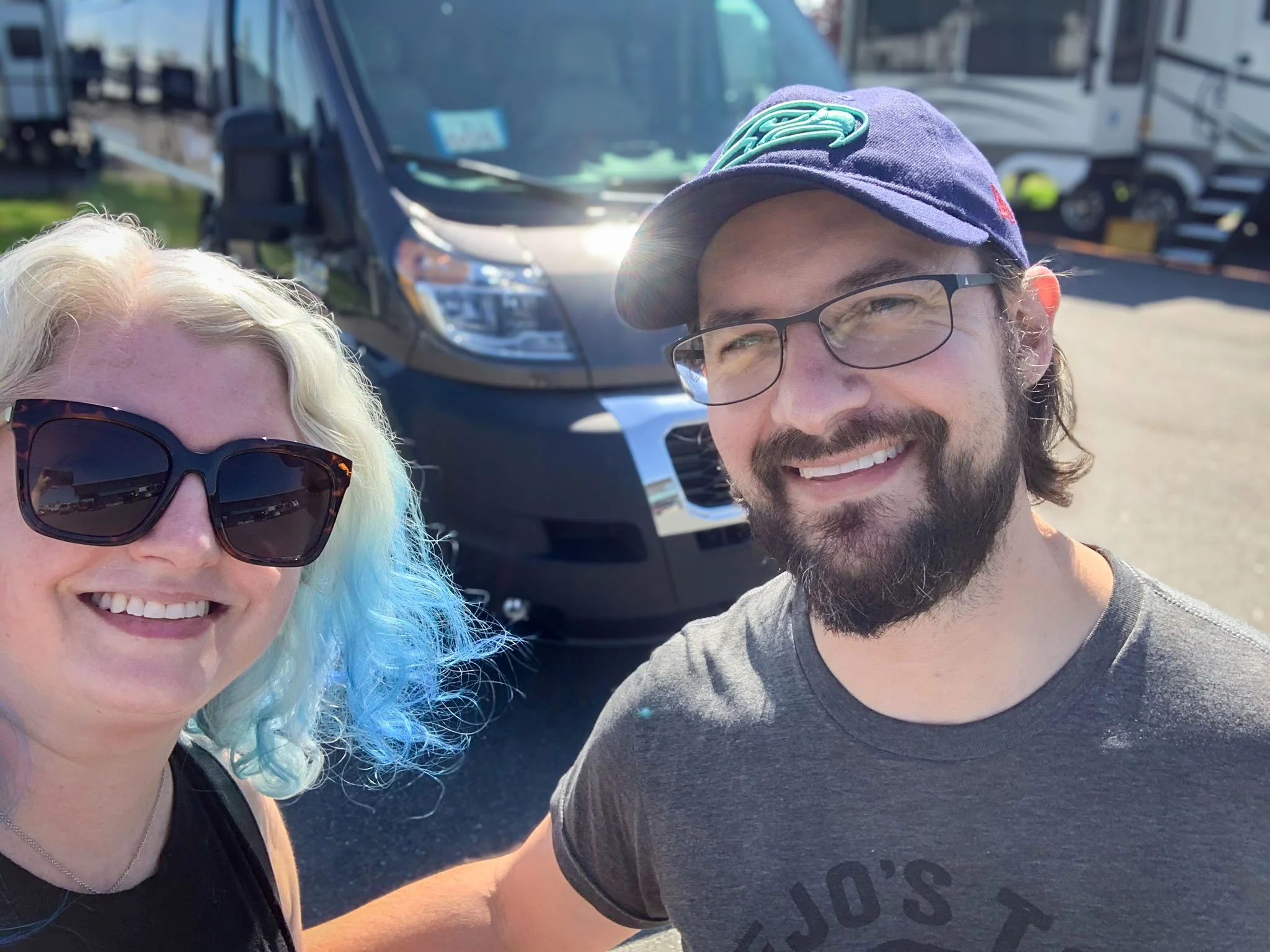 A man and a woman take a selfie photo in front of a dark gray camper van they just purchased in Fife, Washington.