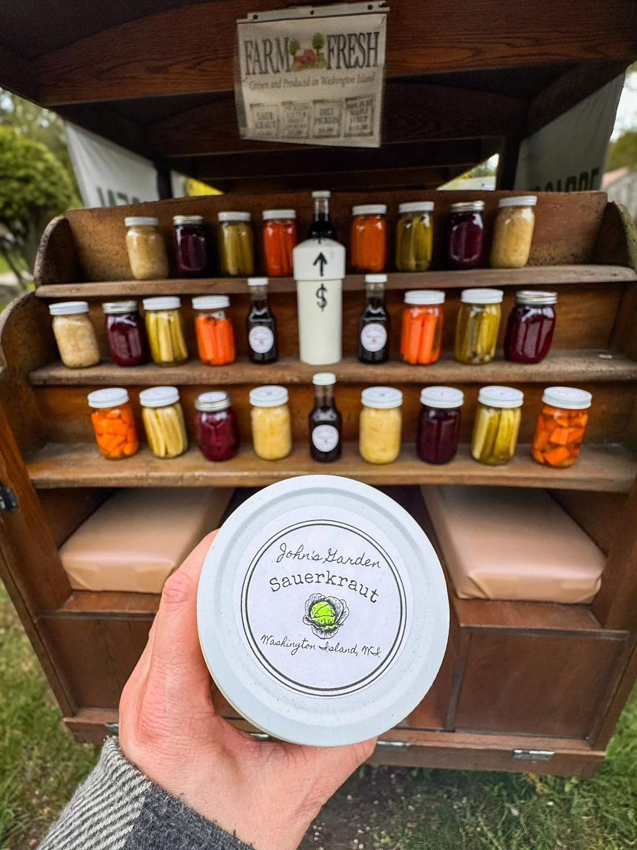 A hand holds a jar of sauerkraut from John's Garden on Washington Island, Wisconsin, with the shelves of the farm stand in the background.