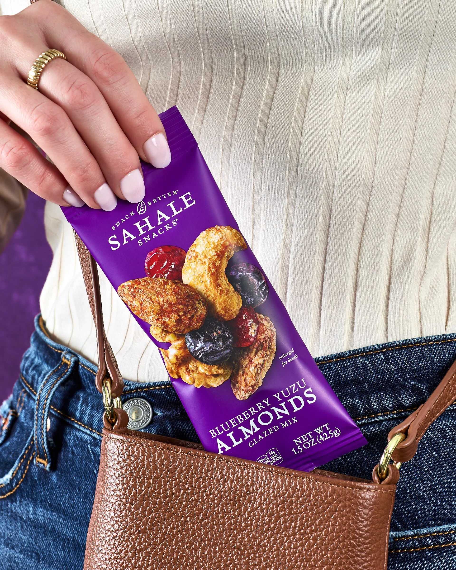 A person in a beige top and blue jeans holding a purple bag of Sahale Snacks Blueberry Yuzu Almonds in place of a pocket, with a tan textured purse hanging from their shoulder.