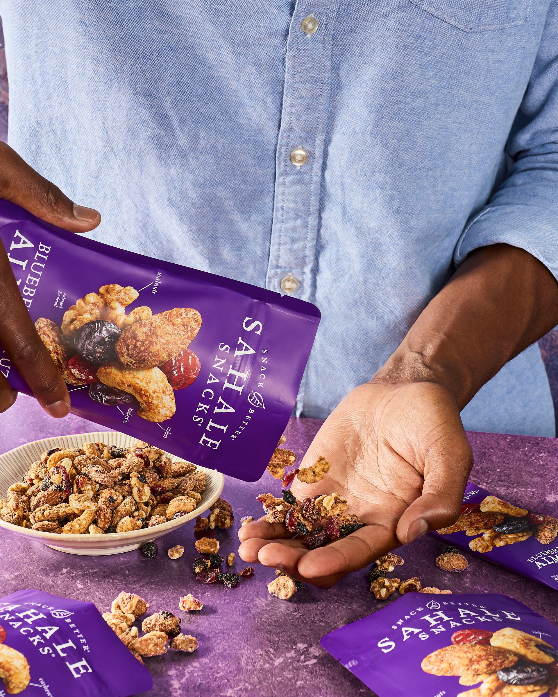 Person pouring blueberry almond snack mix from purple packaging into their hand over a purple surface, with additional snack packets and a bowl of snack mix on the table.