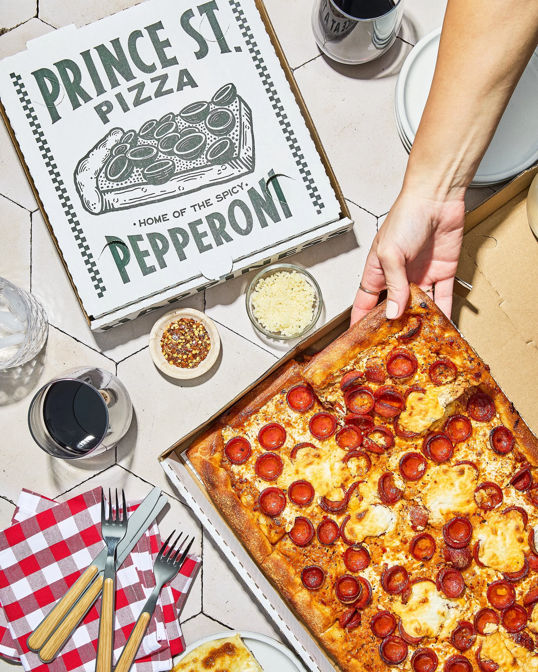 Person lifting a slice of pepperoni pizza from a box at a table with drinks, utensils, and pizza ingredients, with a pizza box labeled "Prince St Pizza Home of the Spicy Pepperoni."