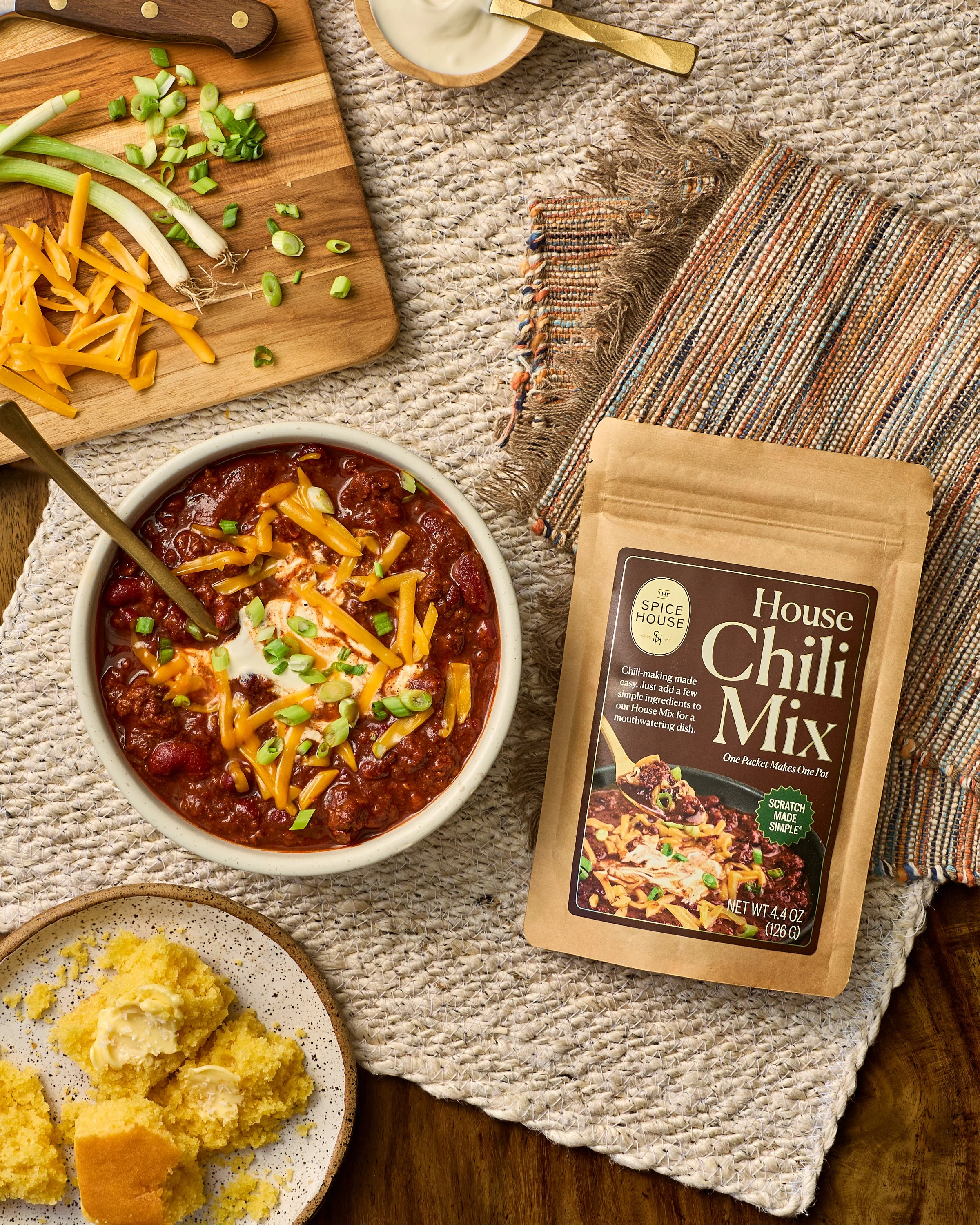 A bowl of chili topped with shredded cheddar cheese and chopped green onions, with a bag of The Spice House chili mix and a plate of cornbread and butter nearby, set on a woven tablecloth. 