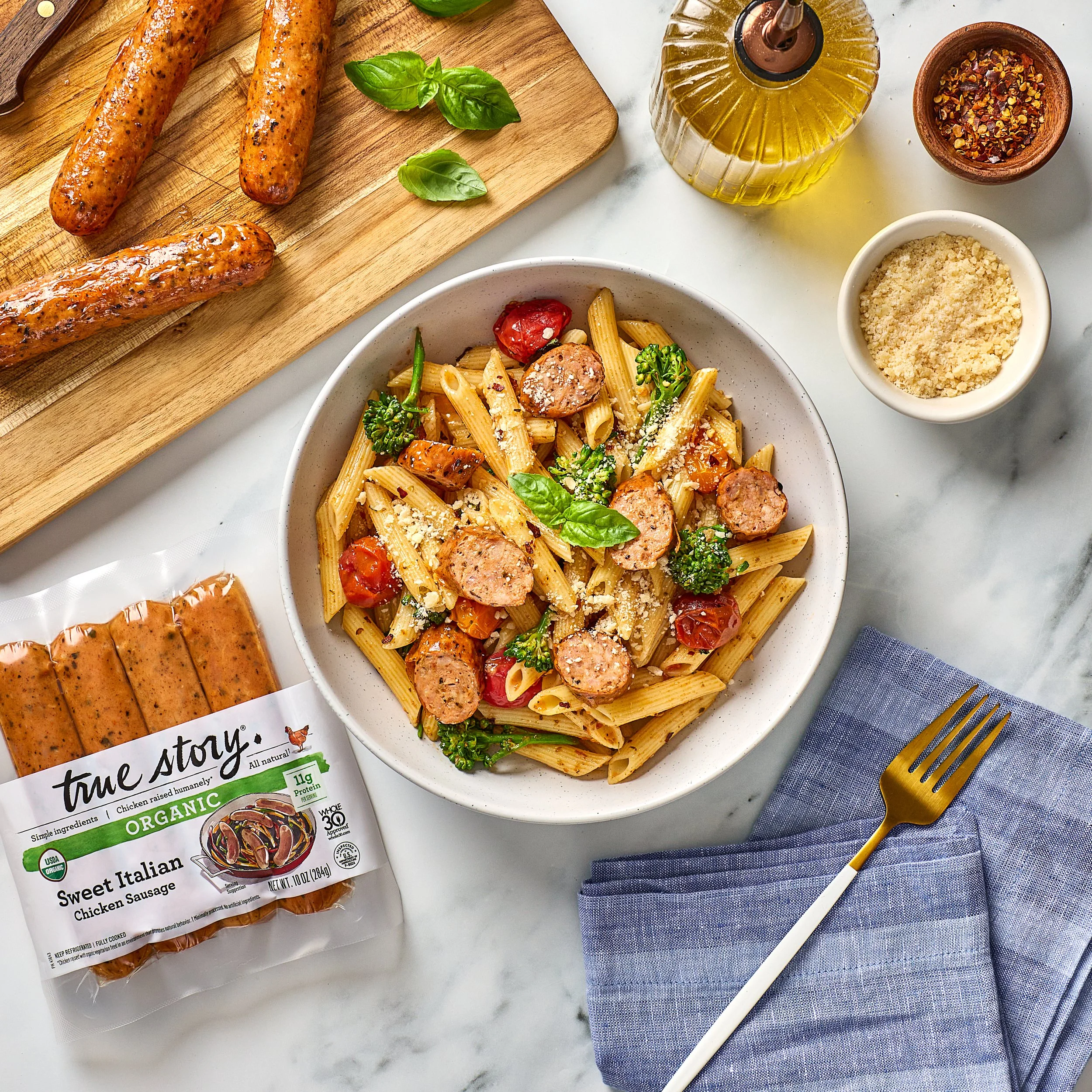 Box of True Story organic chicken sausage, a bowl of pasta with sausage slices and cherry tomatoes, basil leaves, a bottle of olive oil, red pepper flakes, and grated cheese on a marble surface with a blue napkin and yellow chopsticks.
