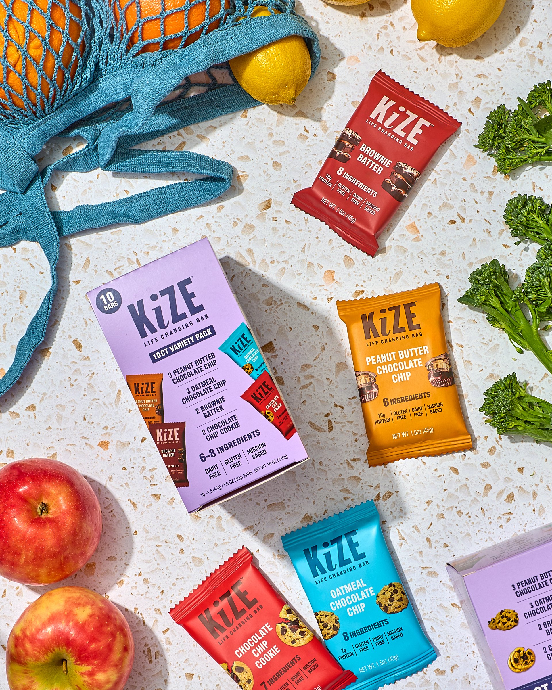 Assorted KiZE snack bars, a purple variety pack box, fresh apples, lemons in a blue net bag, and green kale on a white speckled surface.