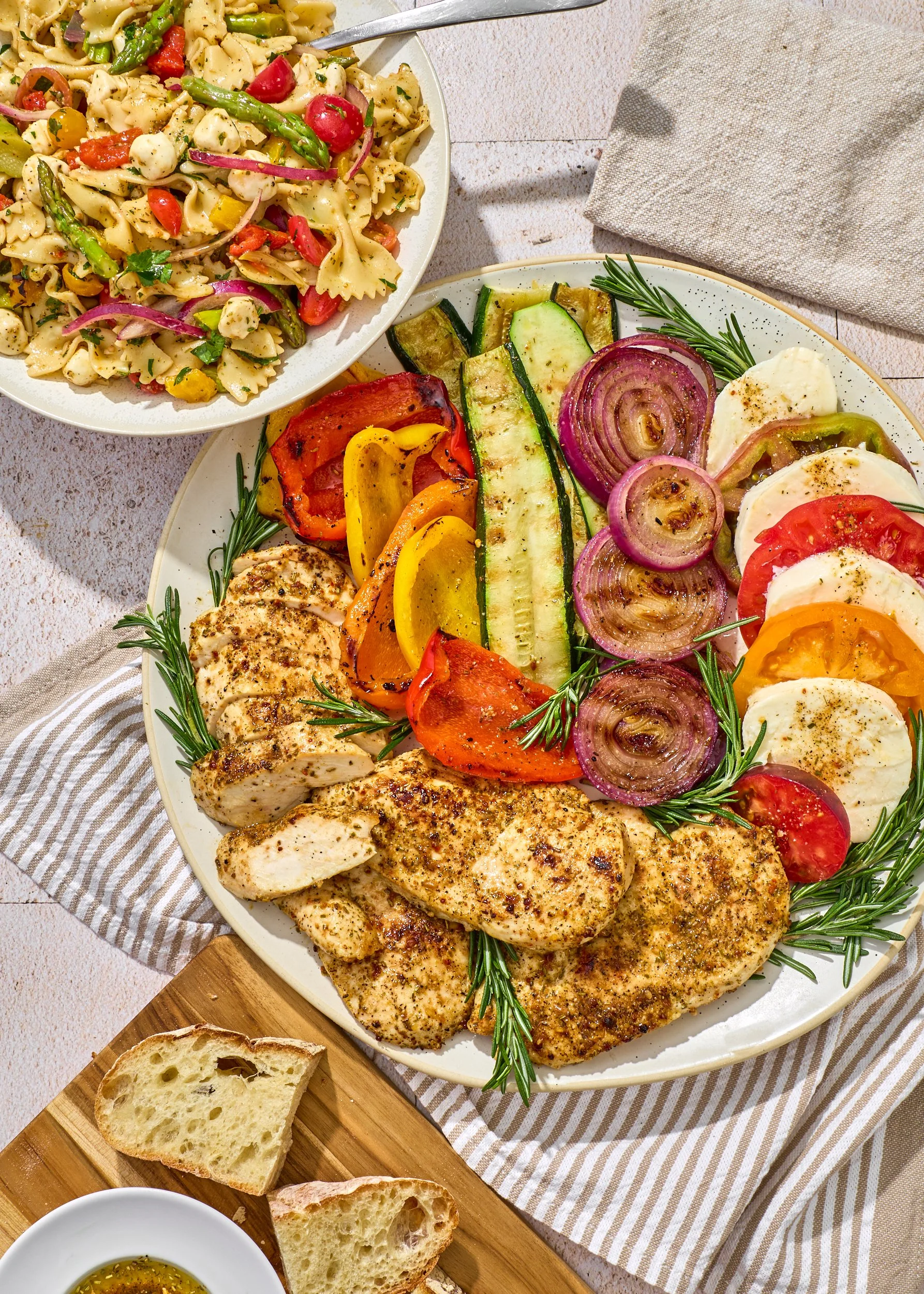 A platter of grilled chicken breast with herbs, served with sliced tomatoes, cucumbers, red onions, and roasted bell peppers, garnished with sprigs of rosemary.
