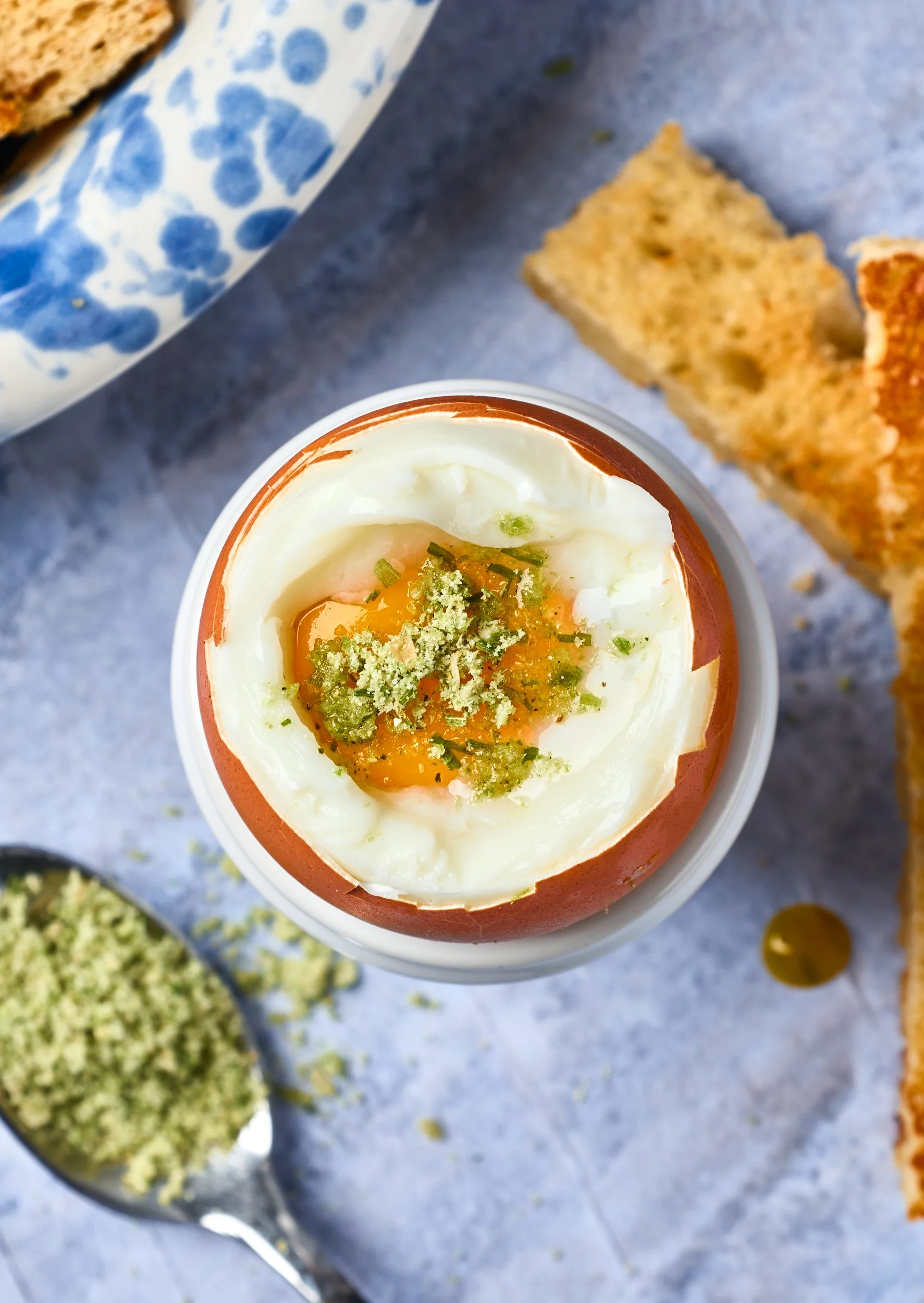 Soft-boiled egg in an egg cup topped with chopped herbs, with toasted bread slices and a spoonful of green seasoning or relish on a light textured surface.