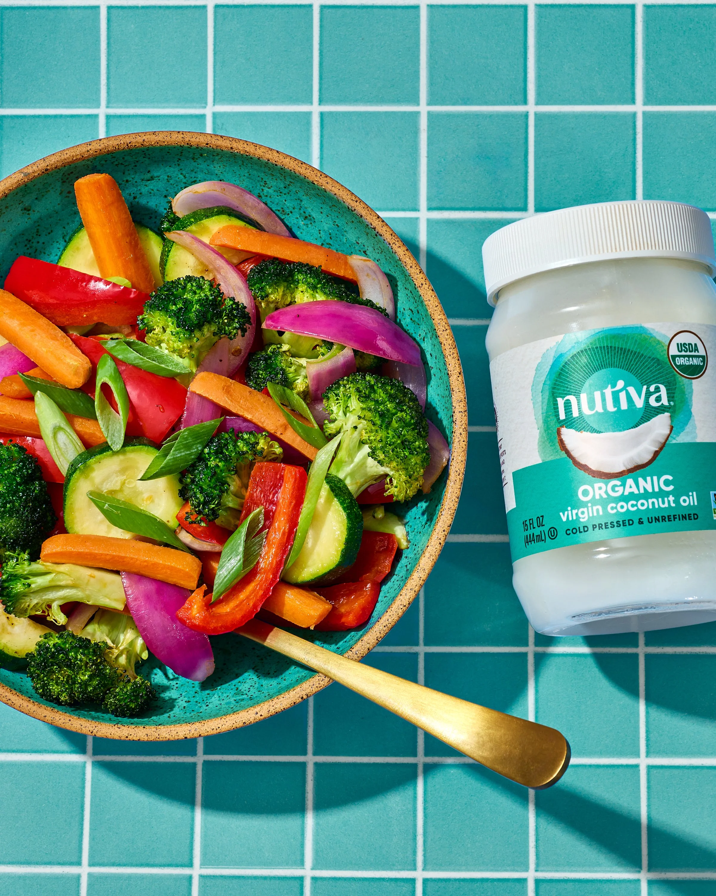 A turquoise bowl filled with sliced red peppers, broccoli, cucumbers, carrots, and onions, with a gold spoon inside. To the right, there is a white container of Nutiva organic virgin coconut oil on a turquoise tiled surface.