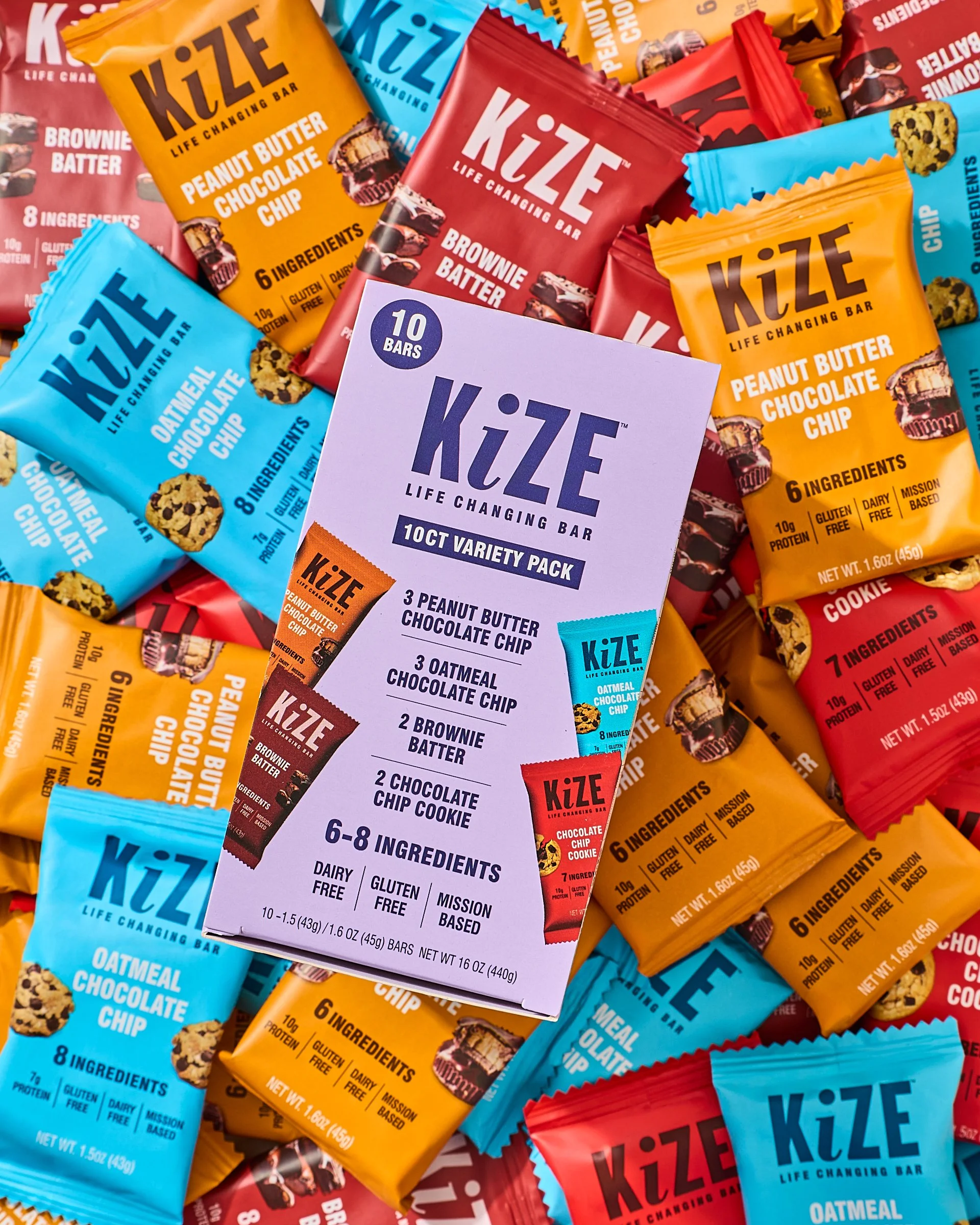 Box labeled "Kize Life Changing Bar 10ct Variety Pack" surrounded by colorful individual snack bars in blue, orange, red, and brown wrappers, with flavors including peanut butter chocolate chip, oatmeal chocolate chip, brownie batter, and chocolate c