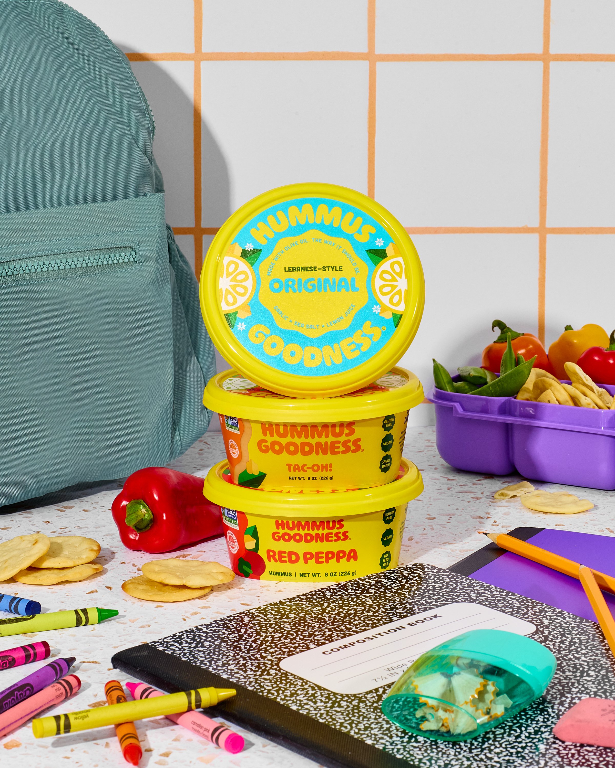 Colorful classroom or workspace with containers of hummus flavor spreads, including original, taco, and red pepper, surrounded by crayons, a notebook, and various school supplies on a white speckled table with a tiled wall in the background.