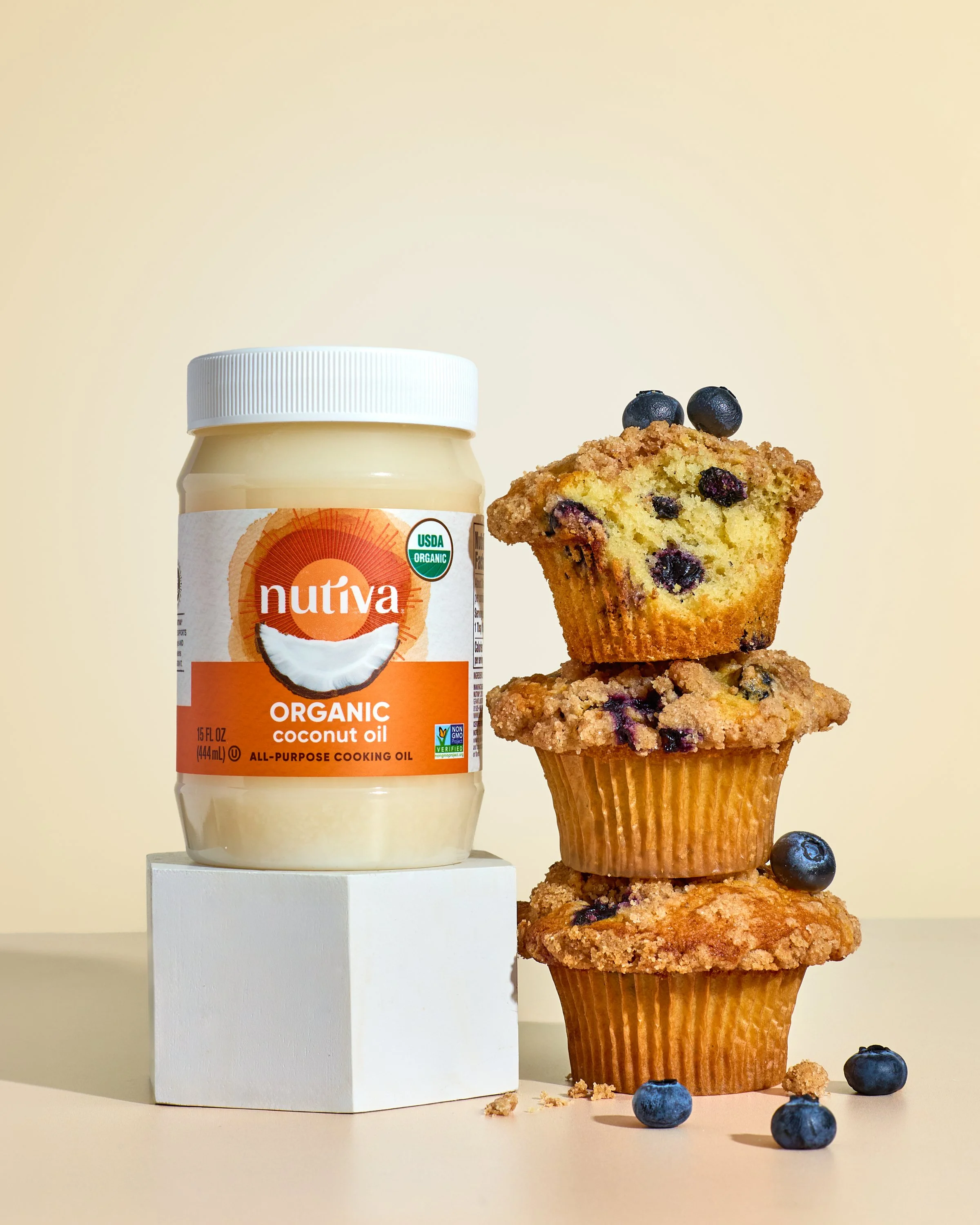 A jar of Nutiva organic coconut oil next to a stack of three blueberry muffins with blueberries on top and blueberries scattered around on a beige surface and a light background.