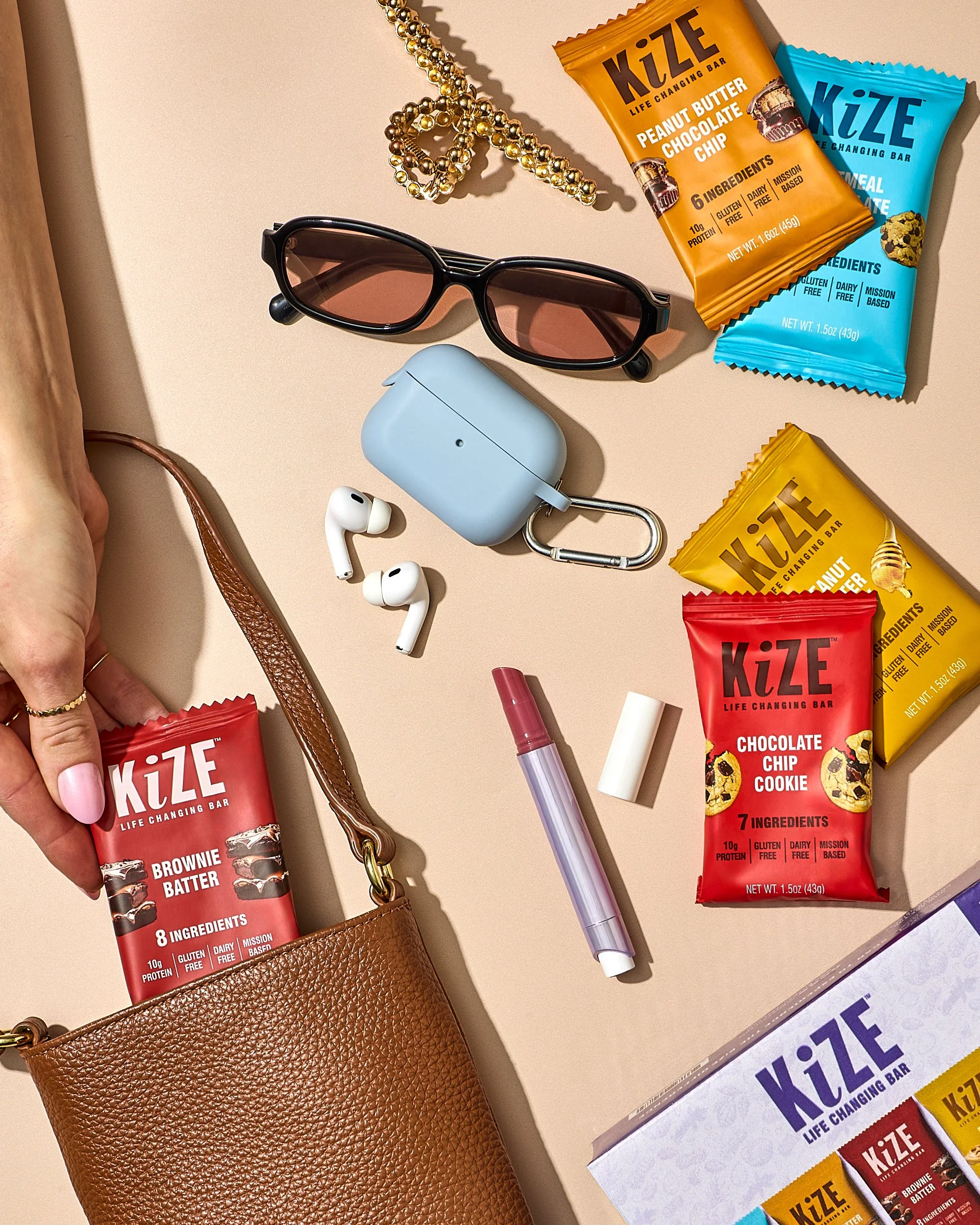 Brown leather purse with metallic hardware on a beige surface. Inside the purse's opening, a hand with an engagement ring and pink nail polish is holding a red package of KiZE Brownie Batter. Items scattered around include blue wireless earbuds with 