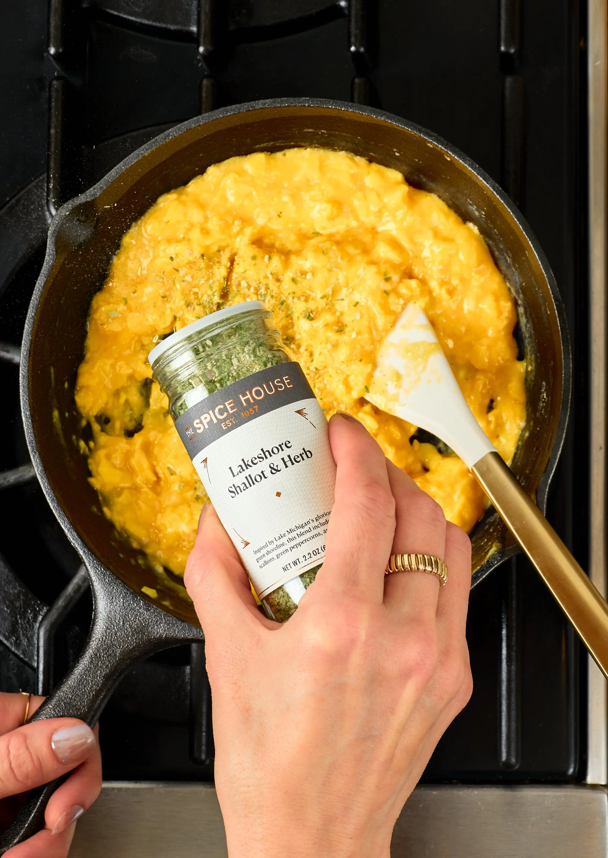 Person adding Lakeshore Shallot & Herb seasoning to scrambled eggs cooking in a skillet, on a stove with a small spatula nearby.