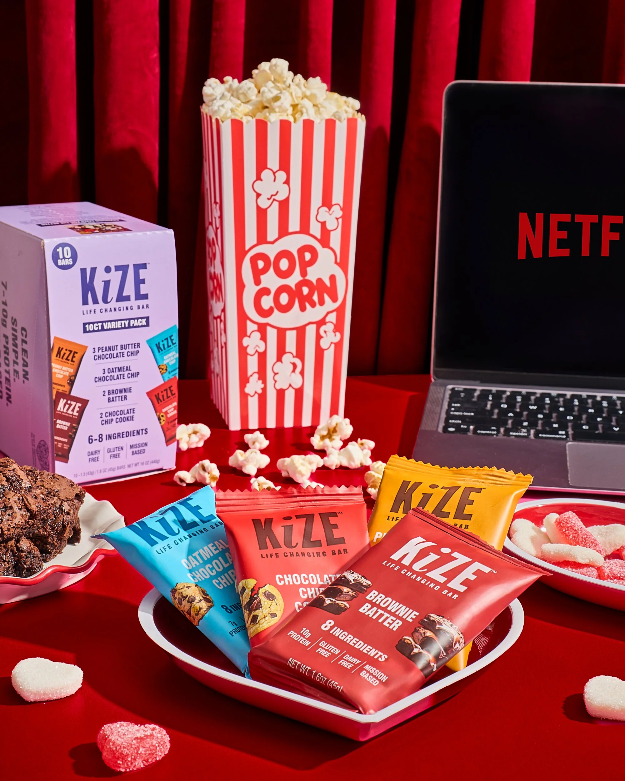 Assorted KiZE meal replacement bars on a red table with popcorn, gummy candies, and a laptop with Netflix logo in the background.