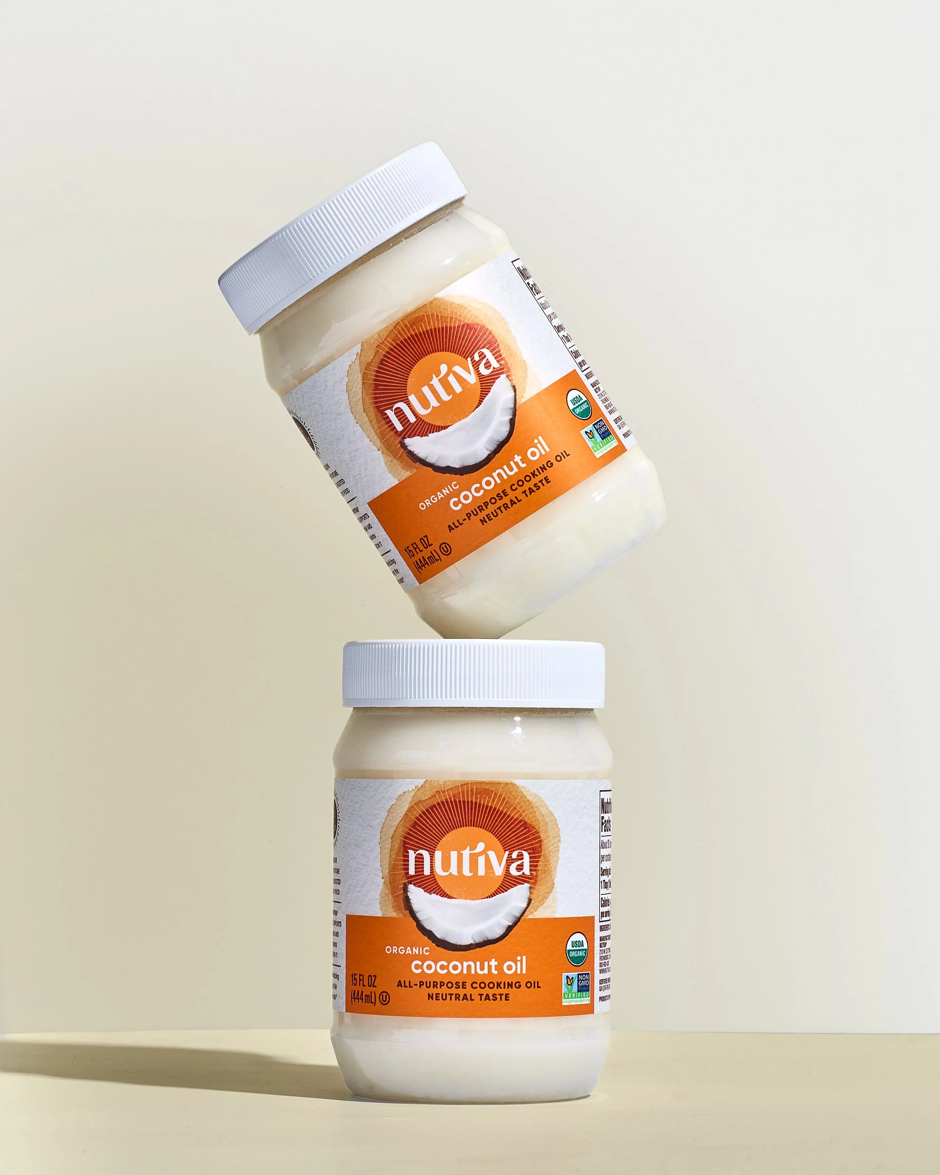 Two containers of Nutiva organic coconut oil, one resting on a surface and the other balanced above it at an angle. The labels are orange and white, featuring a coconut image and the Nutiva logo.