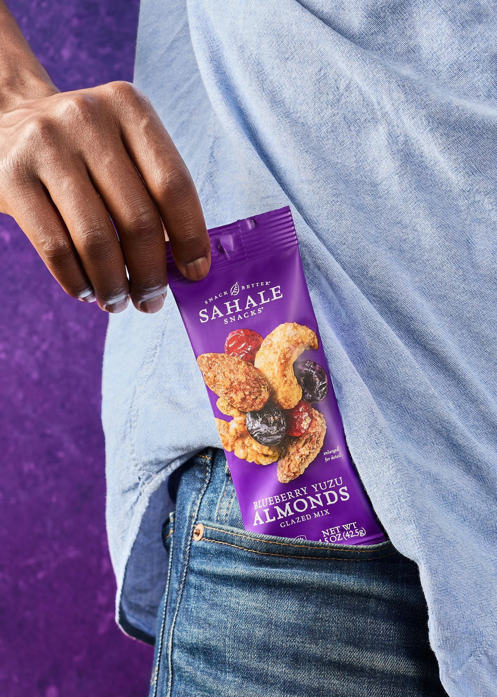 A person is holding a purple pack of Sahale Snacks Blueberry Yuzu Almonds glzed mix, with some whole blueberries and almonds on the packaging, near their jeans pocket.