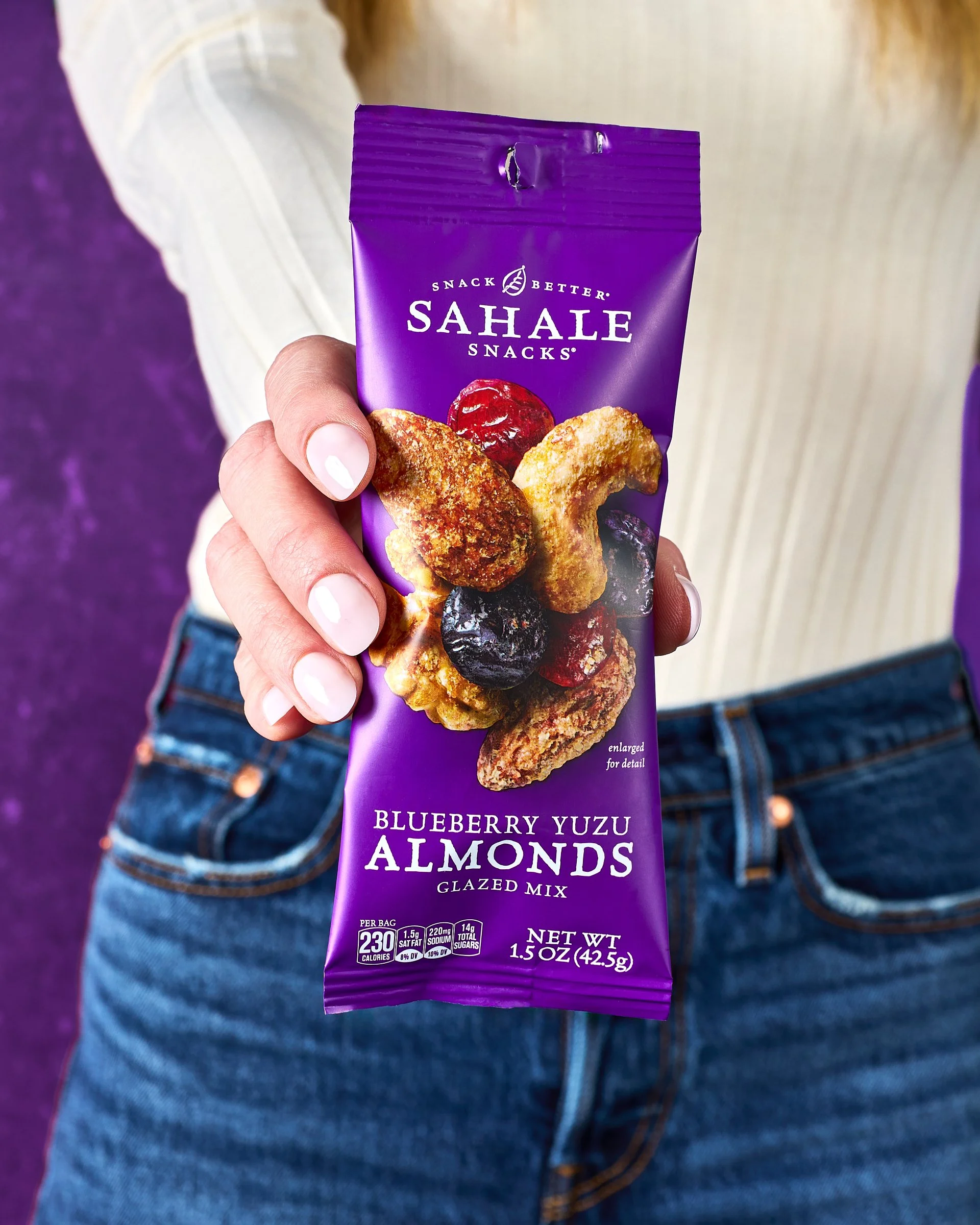 A person holding a purple bag of Sahale Snacks Blueberry Yuzu Almonds Glazed Mix in front of a background with purple and beige colors. The bag has an image of almonds and blueberries on it.
