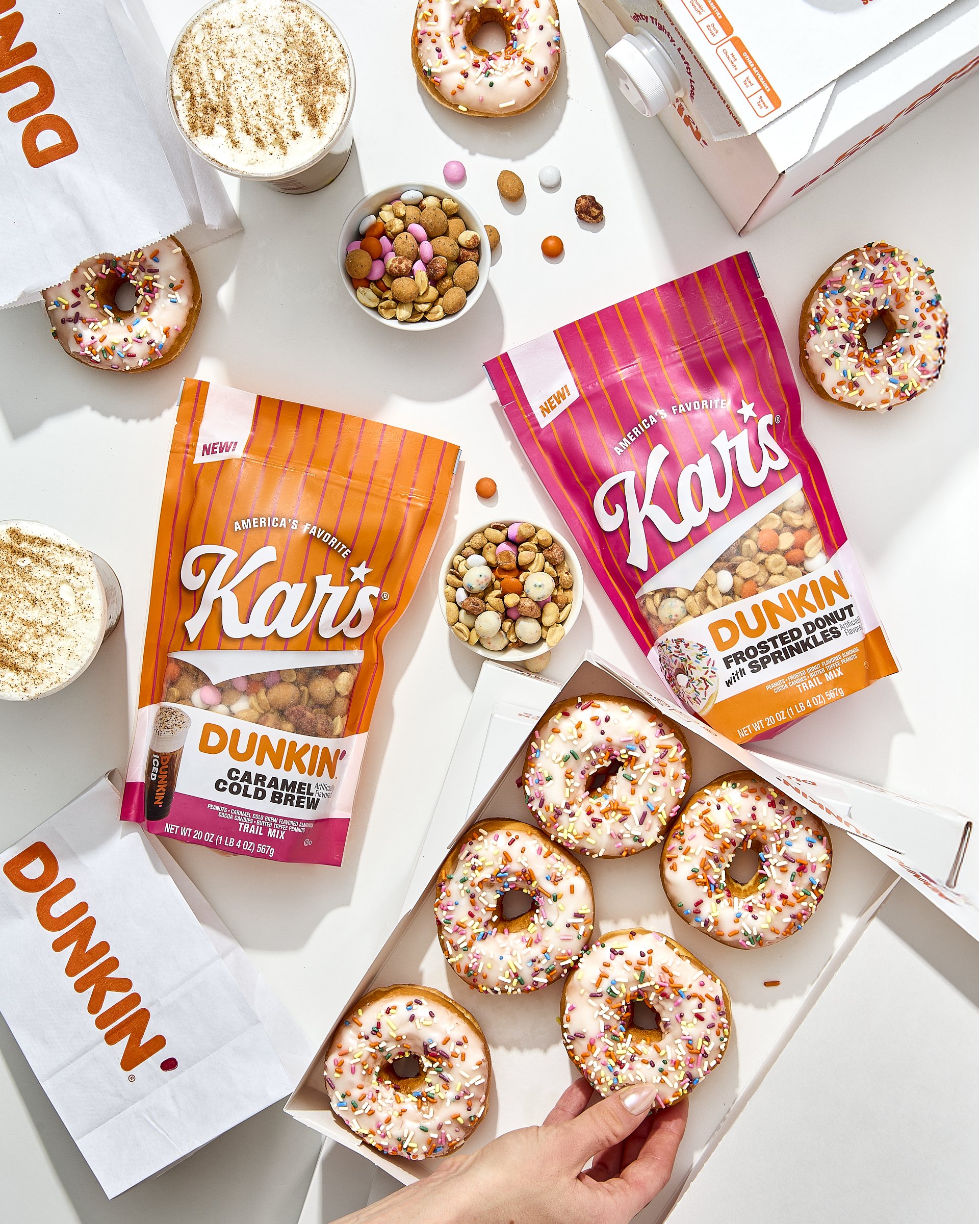 Doughnuts with white icing and colorful sprinkles in an open box, two packages of Kar's snack mix, two cups of coffee with foam, and small bowls of snack mix scattered on a white table.