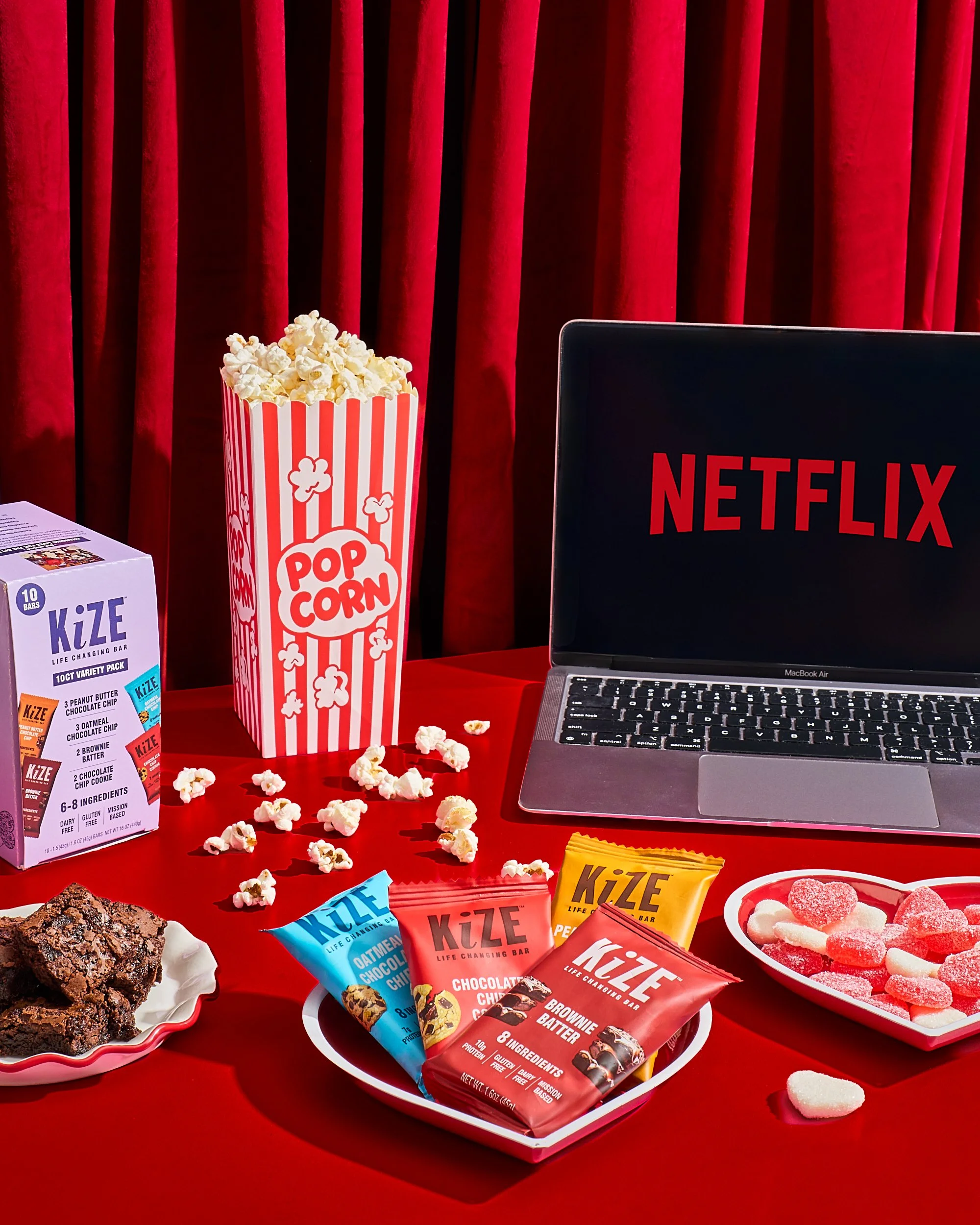 A red table with a laptop displaying the Netflix logo, a large popcorn container, a box of KiZE protein bars, a plate of popcorn with red and white candies, and a small dish of brownies, against red curtains in the background.