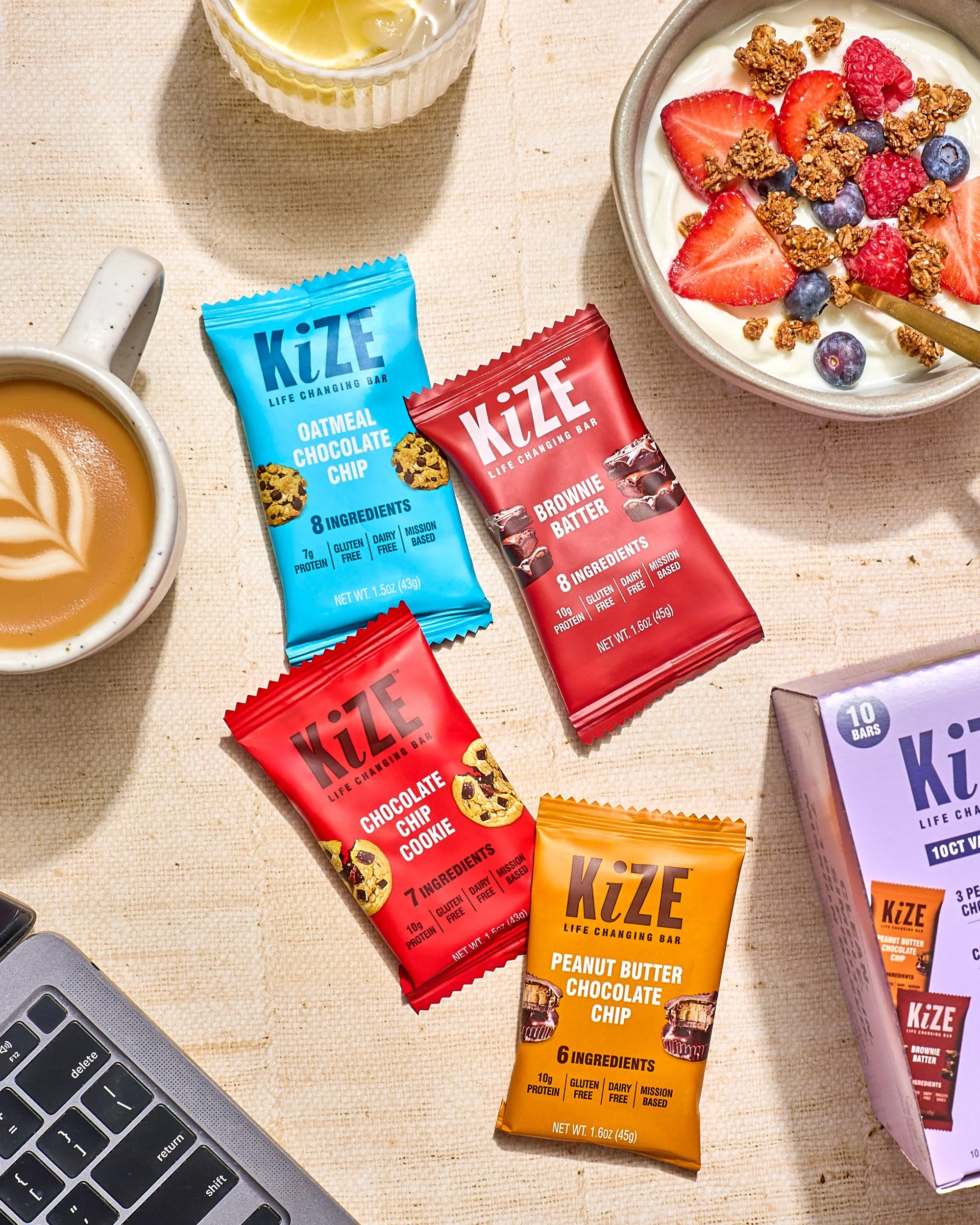 A flat lay of various KIZE protein bars, including oatmeal chocolate chip, brownie batter, chocolate chip cookie, and peanut butter chocolate flavors, with a cup of coffee, a glass of lemon water, and a bowl of yogurt with strawberries, raspberries, 