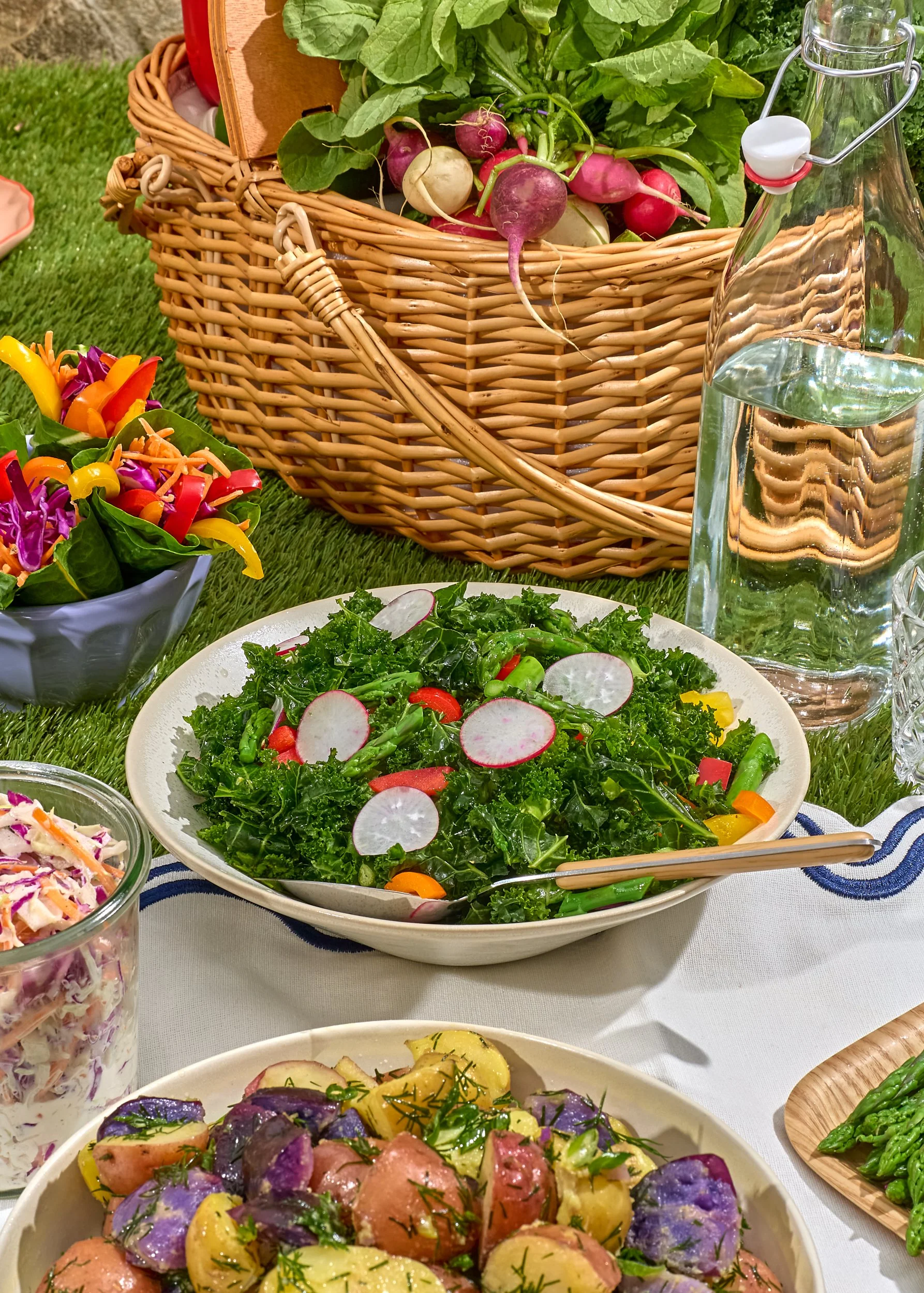 A picnic table set with a basket of colorful radishes, a bowl of mixed salad with radish slices, a plate of roasted potatoes with herbs, a glass of coleslaw, a bowl of colorful bell pepper salad, a clear water bottle, and a glass jar of water, with g