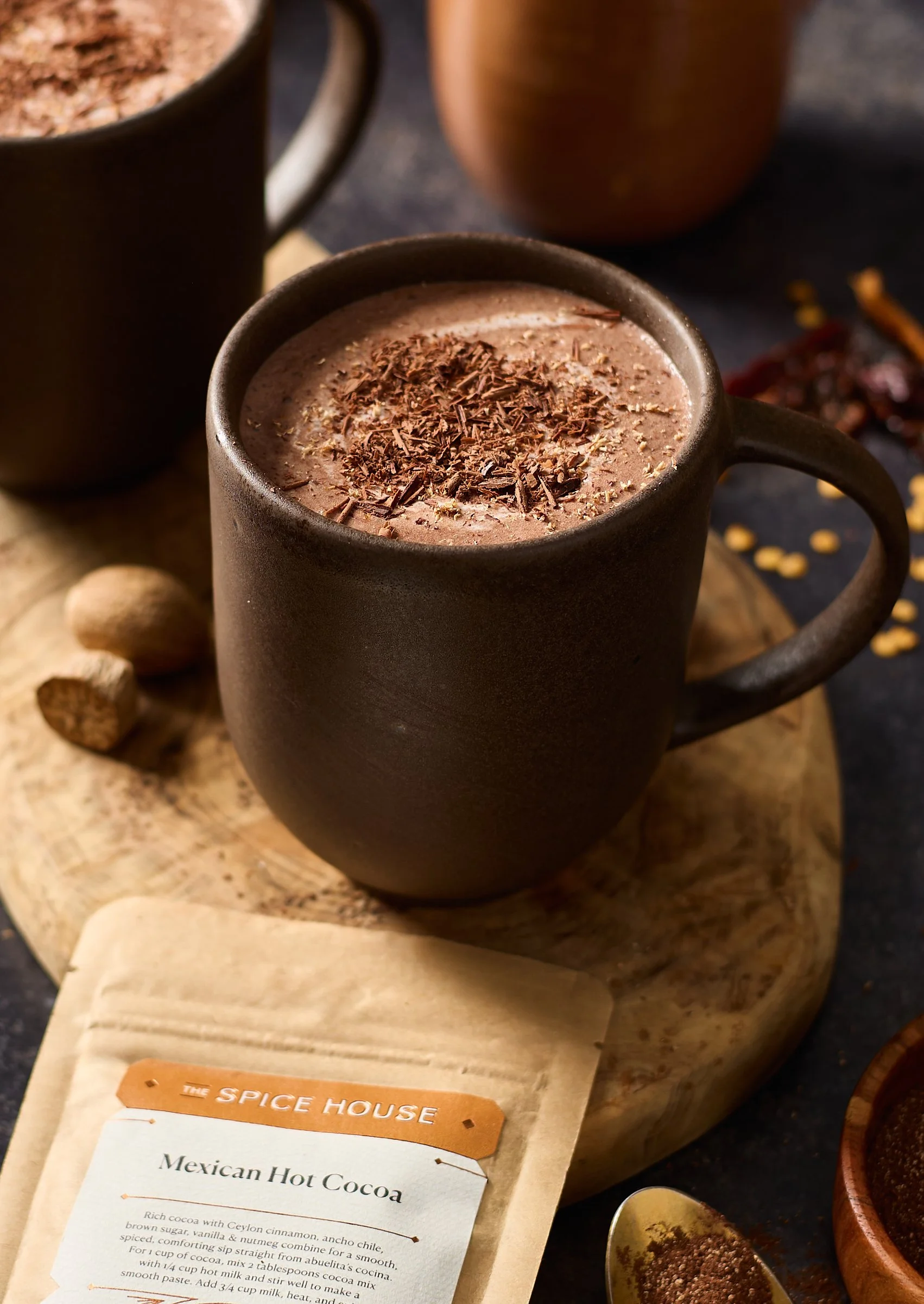 A black ceramic mug filled with Mexican hot cocoa topped with chocolate shavings, placed on a wooden board with a packet of Spice House Mexican Hot Cocoa mix nearby.