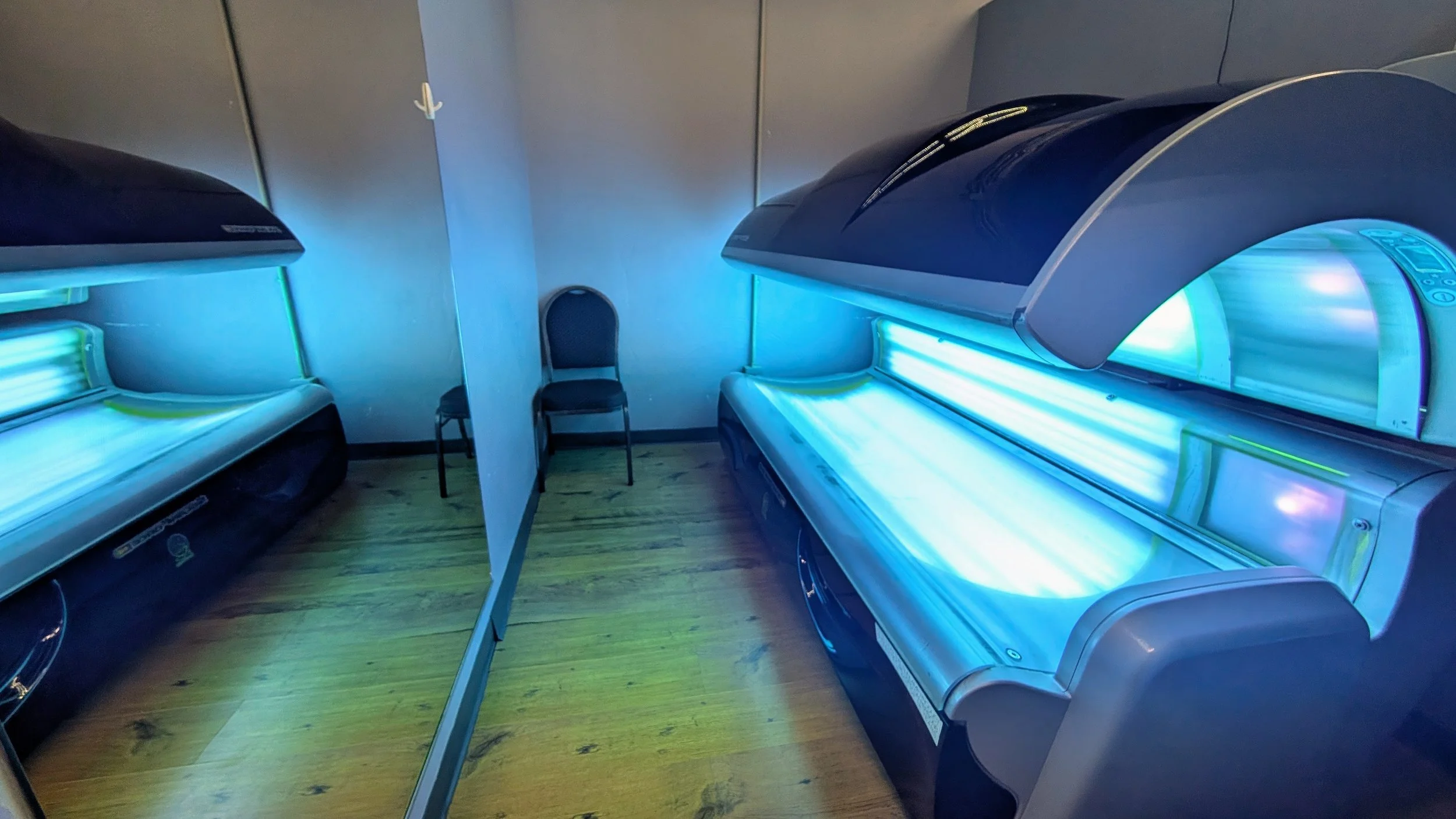 Two tanning beds emitting blue light in a room, separated by a privacy divider, with a single chair against the wall in the background. at shape fitness midwest city