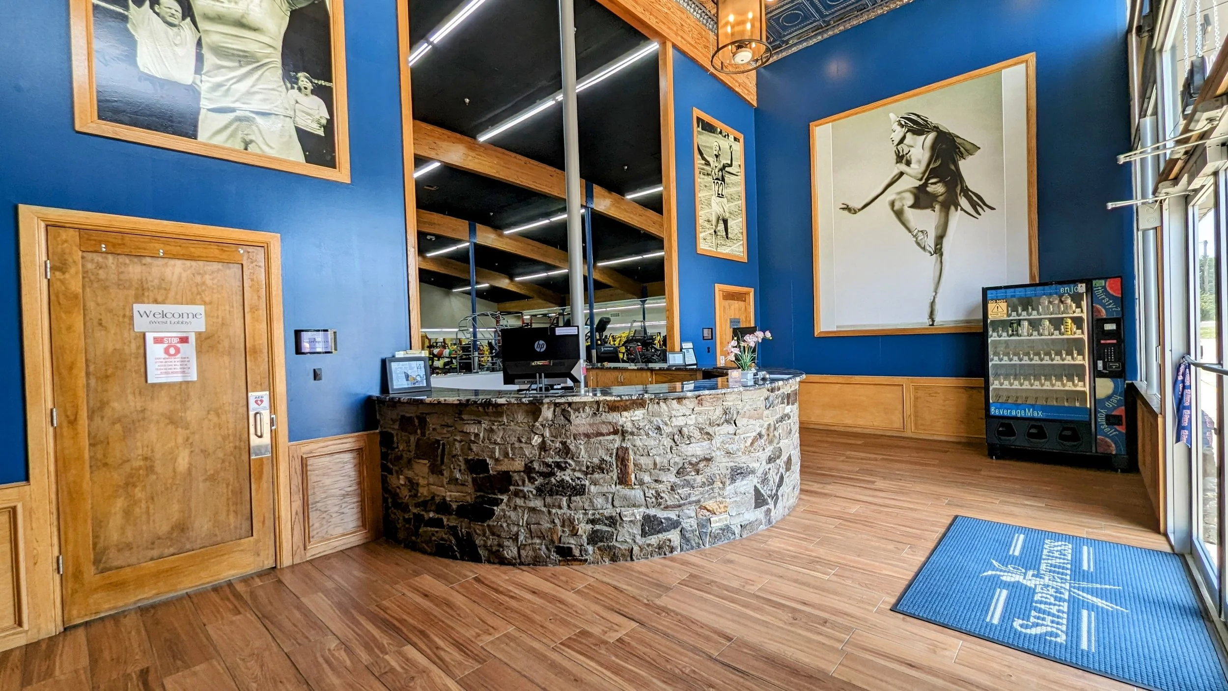 Indoor gym reception area with a curved stone counter, blue walls decorated with large black and white photos, wooden accents, a vending machine, and a welcome sign on the door to the left. Shape fitness El Reno native heritage