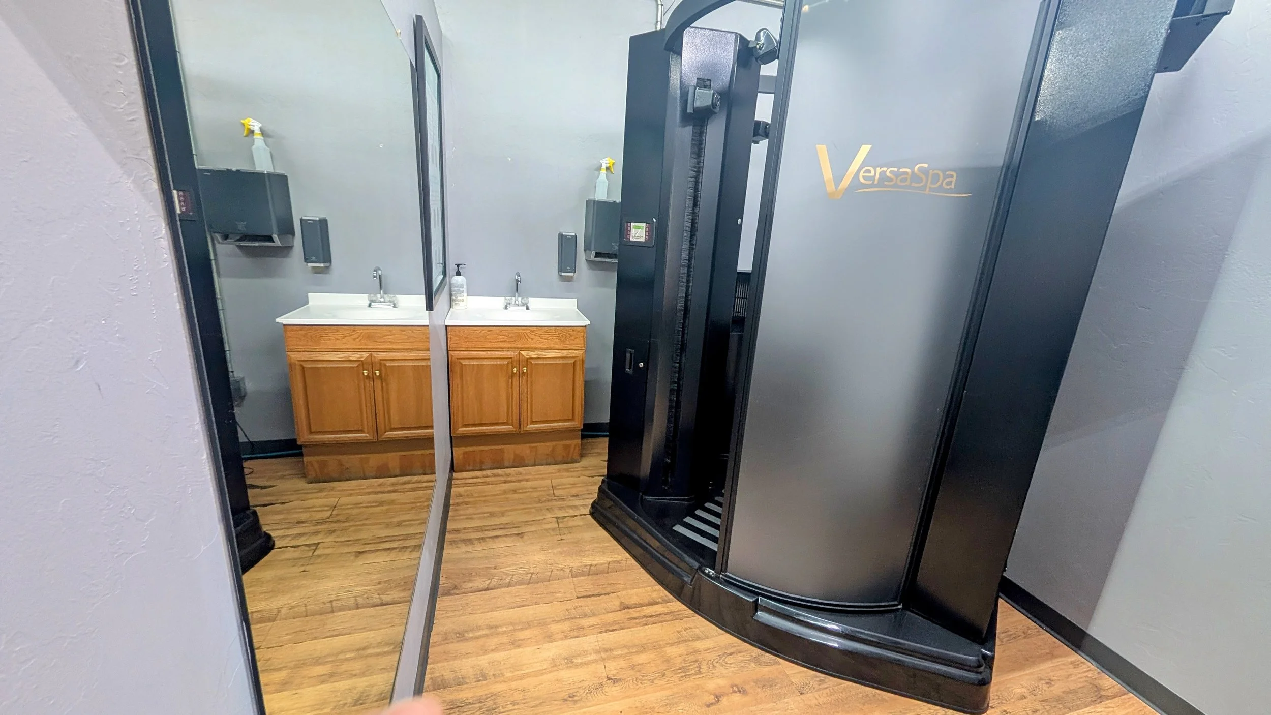 Room with a large black VersaSpa spray tanning booth, mirror reflecting a sink with two faucets, soap dispenser, and brown wooden cabinets, and a wall with spray bottles. at shape fitness midwest city
