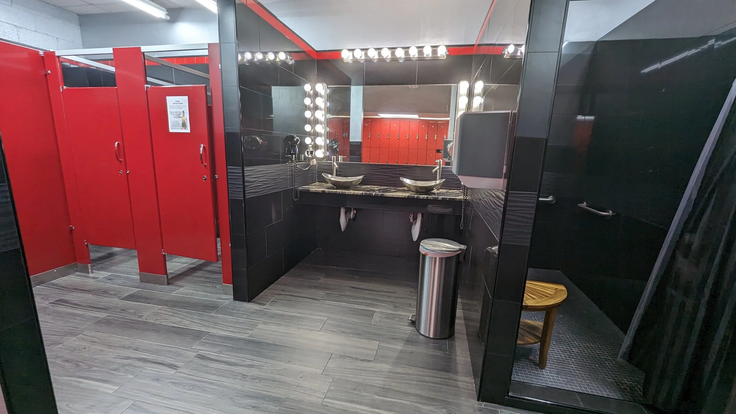 A modern women's restroom with black and red decor, a large mirror with lights, two vessel sinks on a marble countertop, red partition walls, a small wooden stool in a shower stall, and a trash can.Shape fitness El Reno native herita wEST okc okLAhom