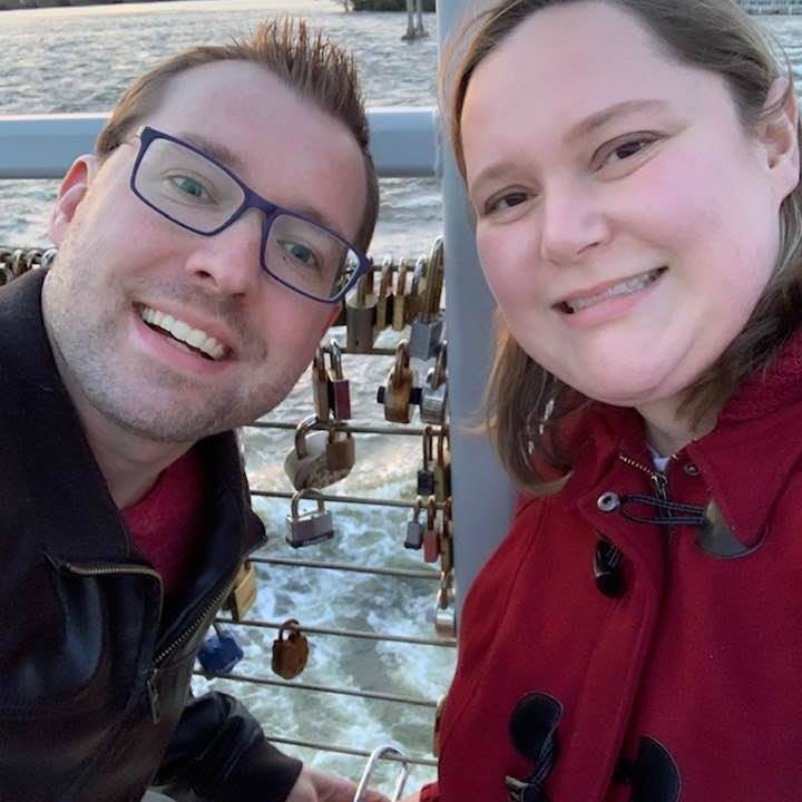 A selfie of a smiling man with glasses and a woman next to him, both on a boat near a body of water with numerous lock padlocks attached to the railing.